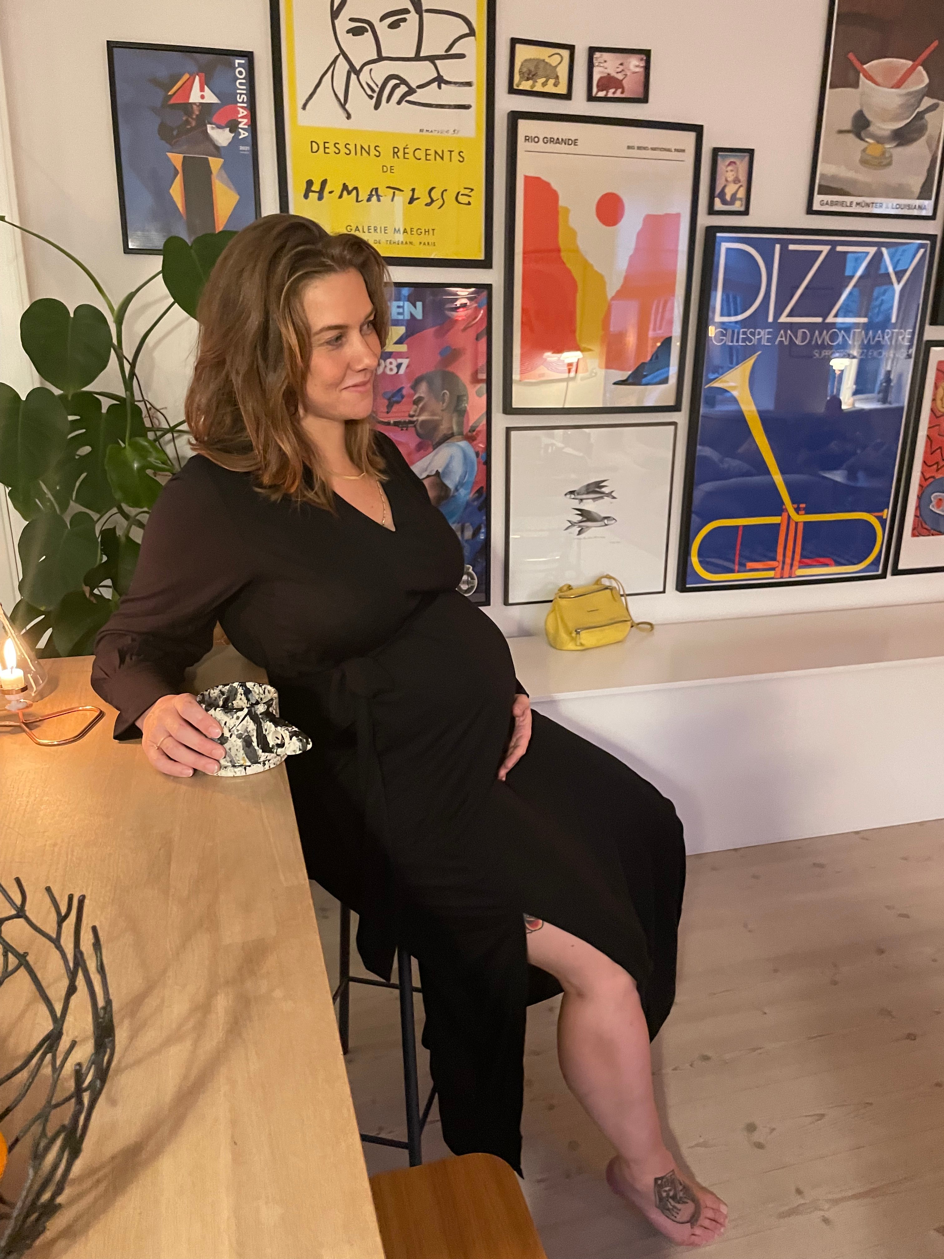 A model is sitting comfortably on a barstool, wearing the AWAN Long Sleeve Kaftan Dress in Warm Black, surrounded by colorful framed posters and art.