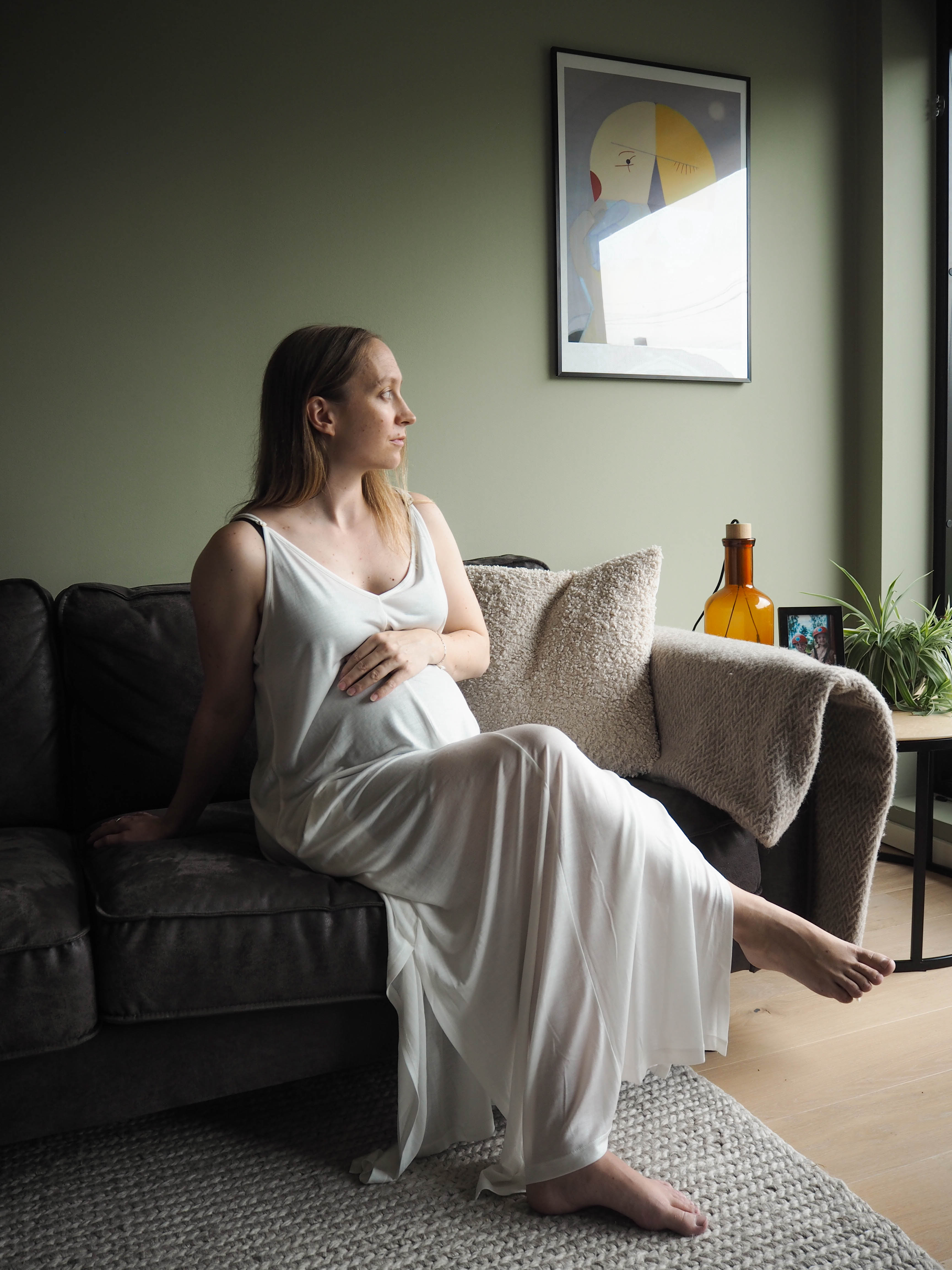 A model is relaxing on a sofa, dressed in the AWAN Boho Slip Dress in Natural White, gazing out the window with a thoughtful look.
