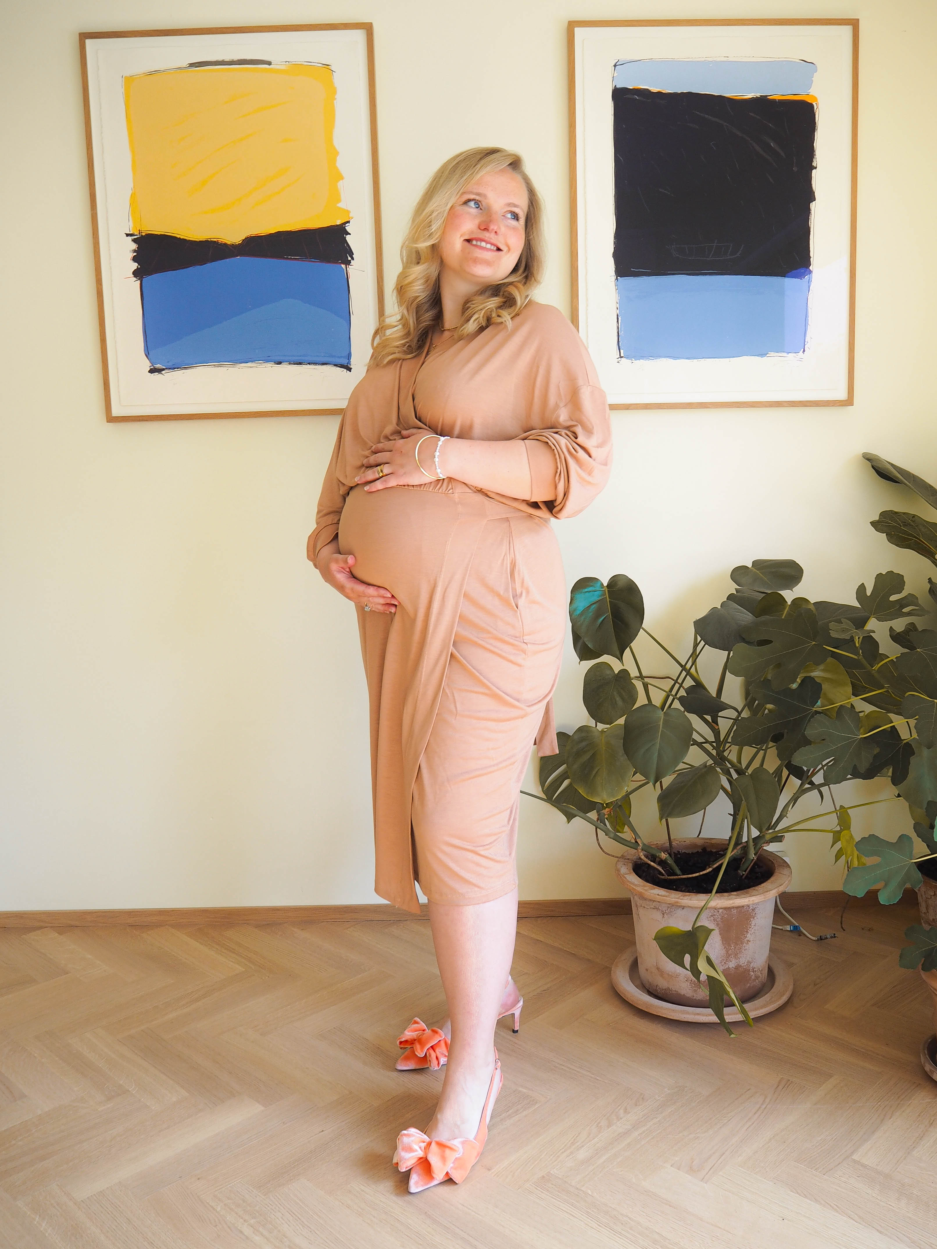 A model stands gracefully in the AWAN Wrap Dress in Camel Rose, posing in front of abstract art pieces with a happy smile and cradling her baby bump.