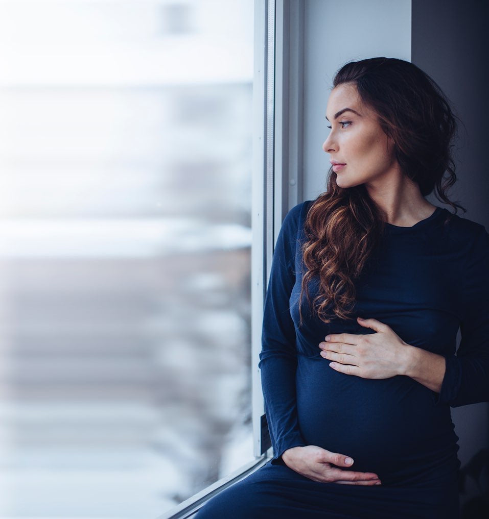 A model is standing by a window, dressed in the AWAN Midnight Dress in Midnight Blue, with one hand gently resting on her baby bump as she gazes outside.