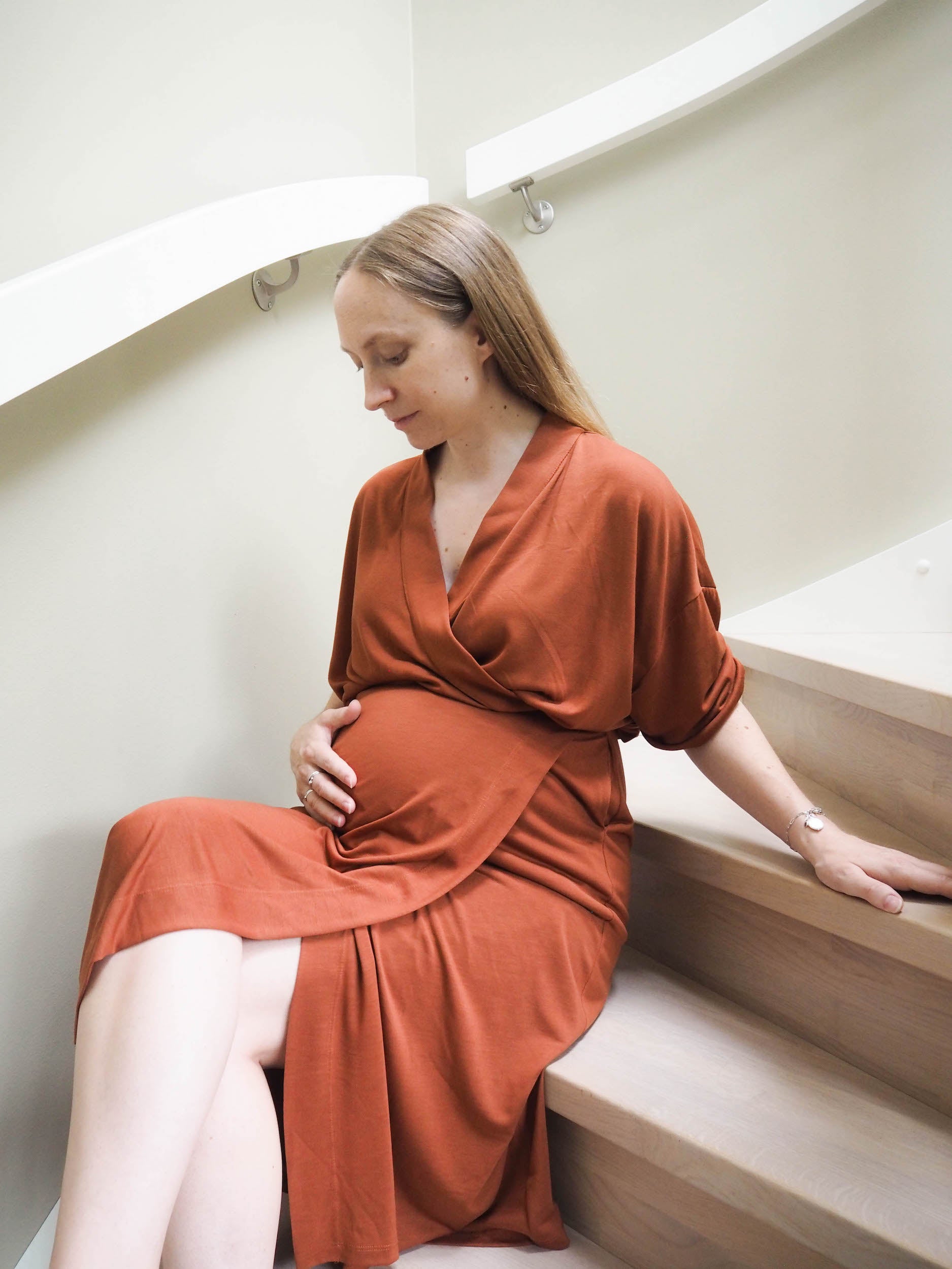 AWAN model showcasing a maternity wrap dress in deep rust, sitting gracefully on a staircase, cradling her belly.