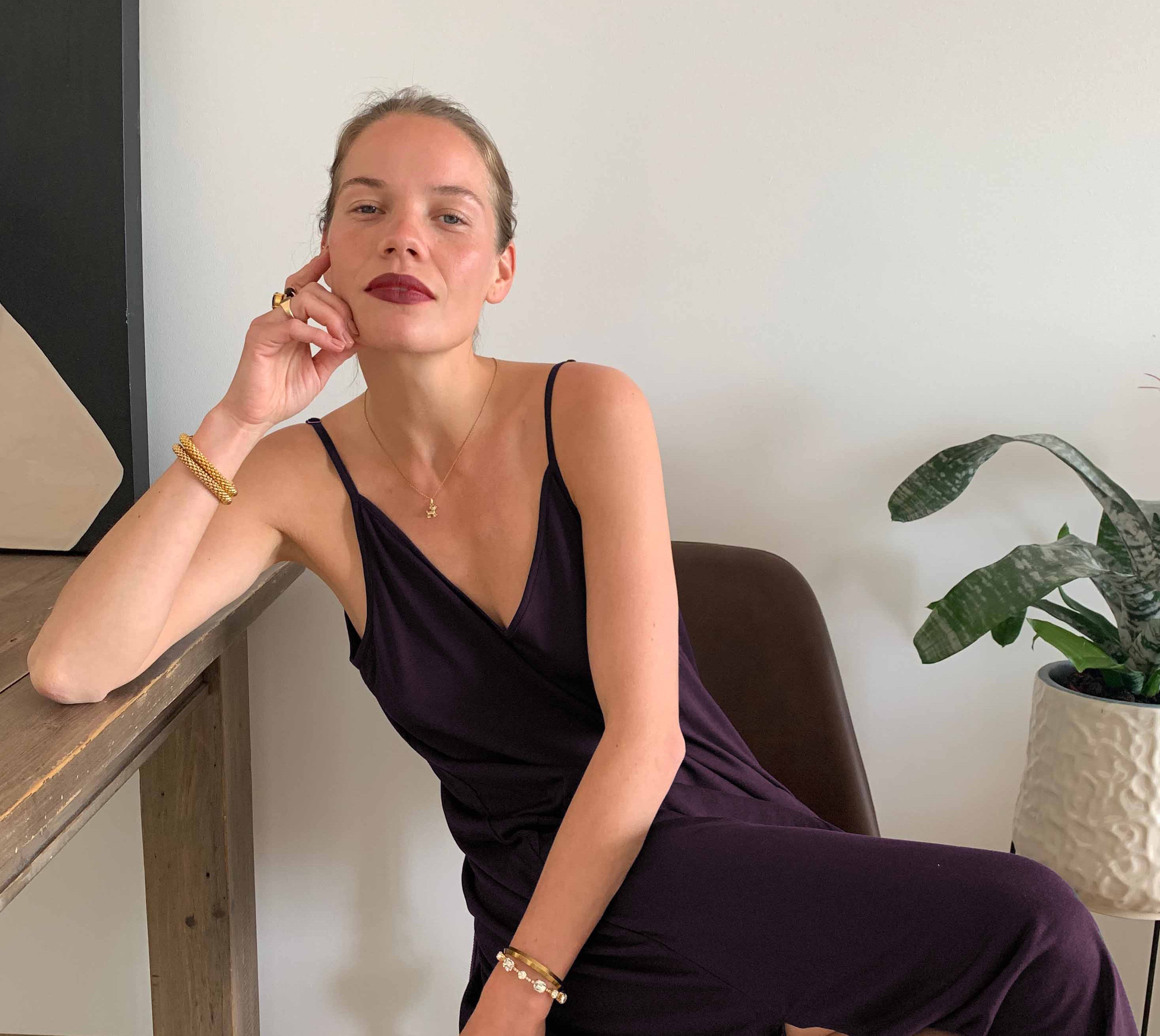 A woman is seated at a wooden table, leaning her elbow on the surface and resting her head on her hand. She is wearing a deep purple spaghetti-strap AWAN boho slip dress in deep plum. She has minimalistic jewelry, including a gold necklace, bracelets, and rings. Her hair is pulled back, and she is looking confidently into the camera with a relaxed expression. A potted plant is visible in the background.