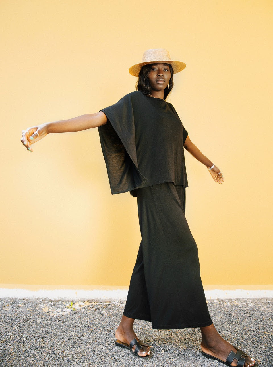 A model poses against a yellow background, wearing a loose-fitting AWAN kaftan top and flow pants in olive green. She pairs the outfit with a straw hat and black sandals, showcasing a relaxed and stylish look.