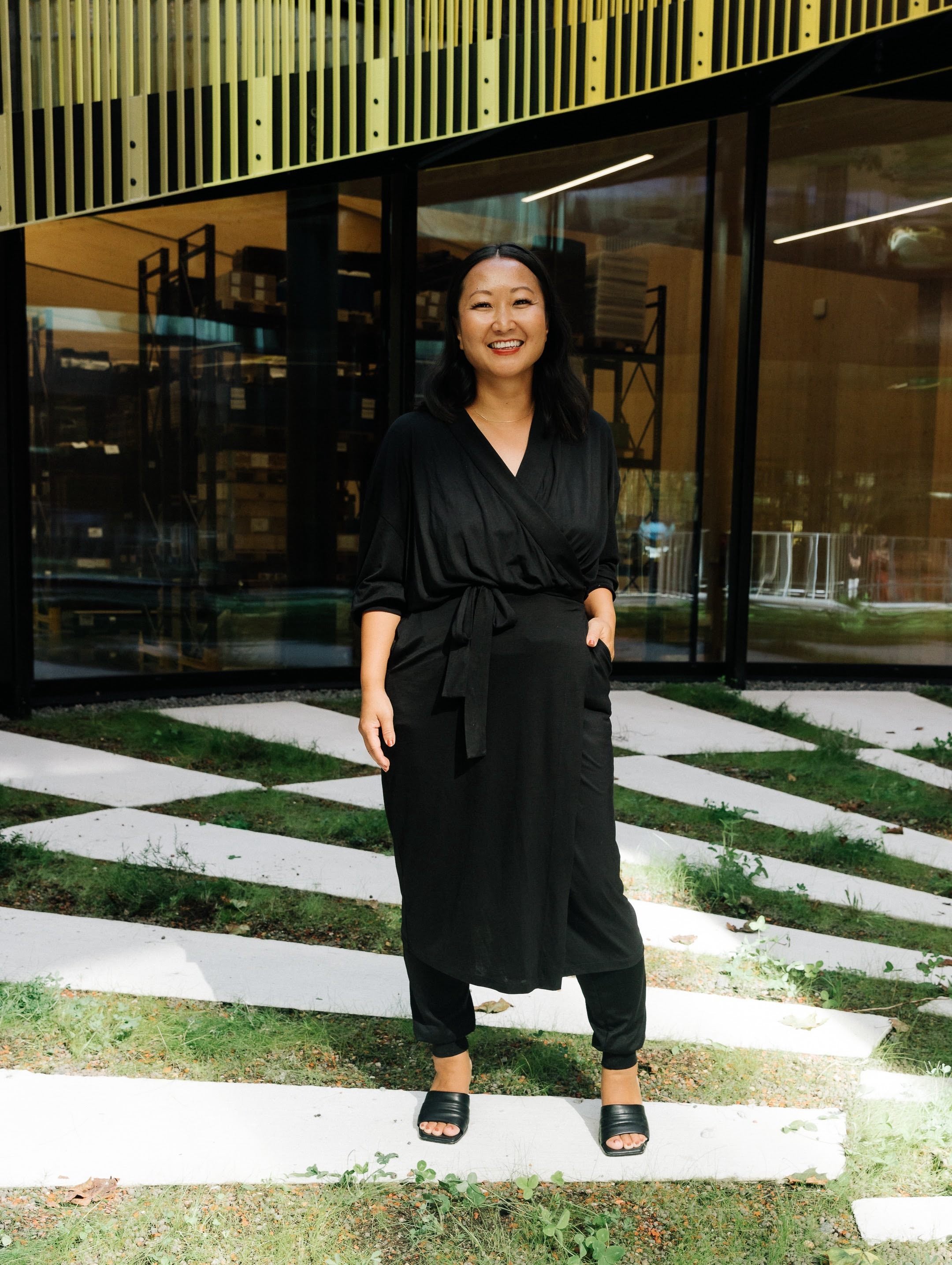 A smiling woman stands outside a modern building, wearing AWAN wrap dress and cuffed pants in warm black. Her outfit exudes confidence and elegance, perfect for a casual yet sophisticated style.