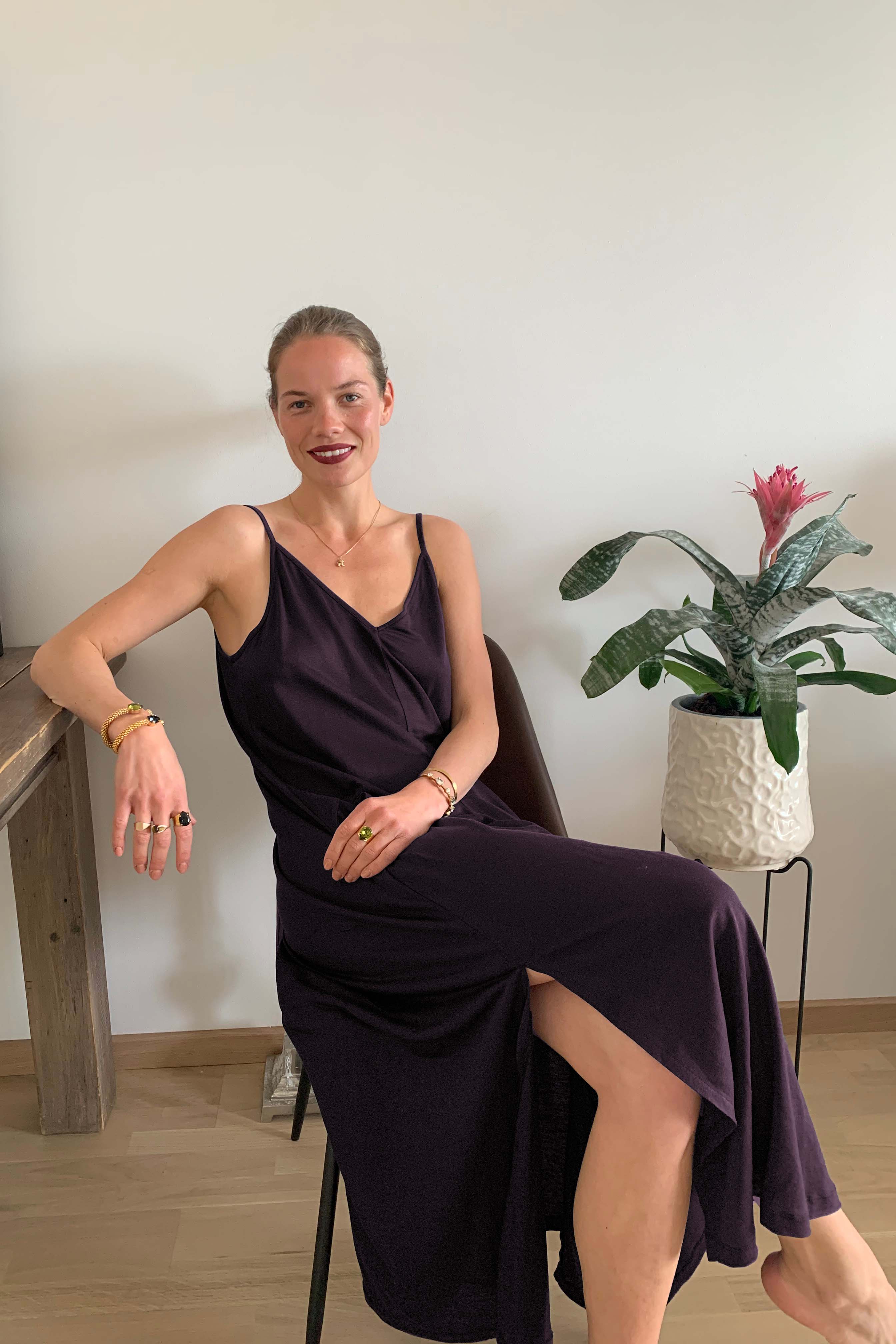 A woman sits gracefully on a chair, wearing AWAN boho slip dress in deep plum. The dress features thin straps and a deep neckline, complemented by gold jewelry, creating a chic and elegant appearance.