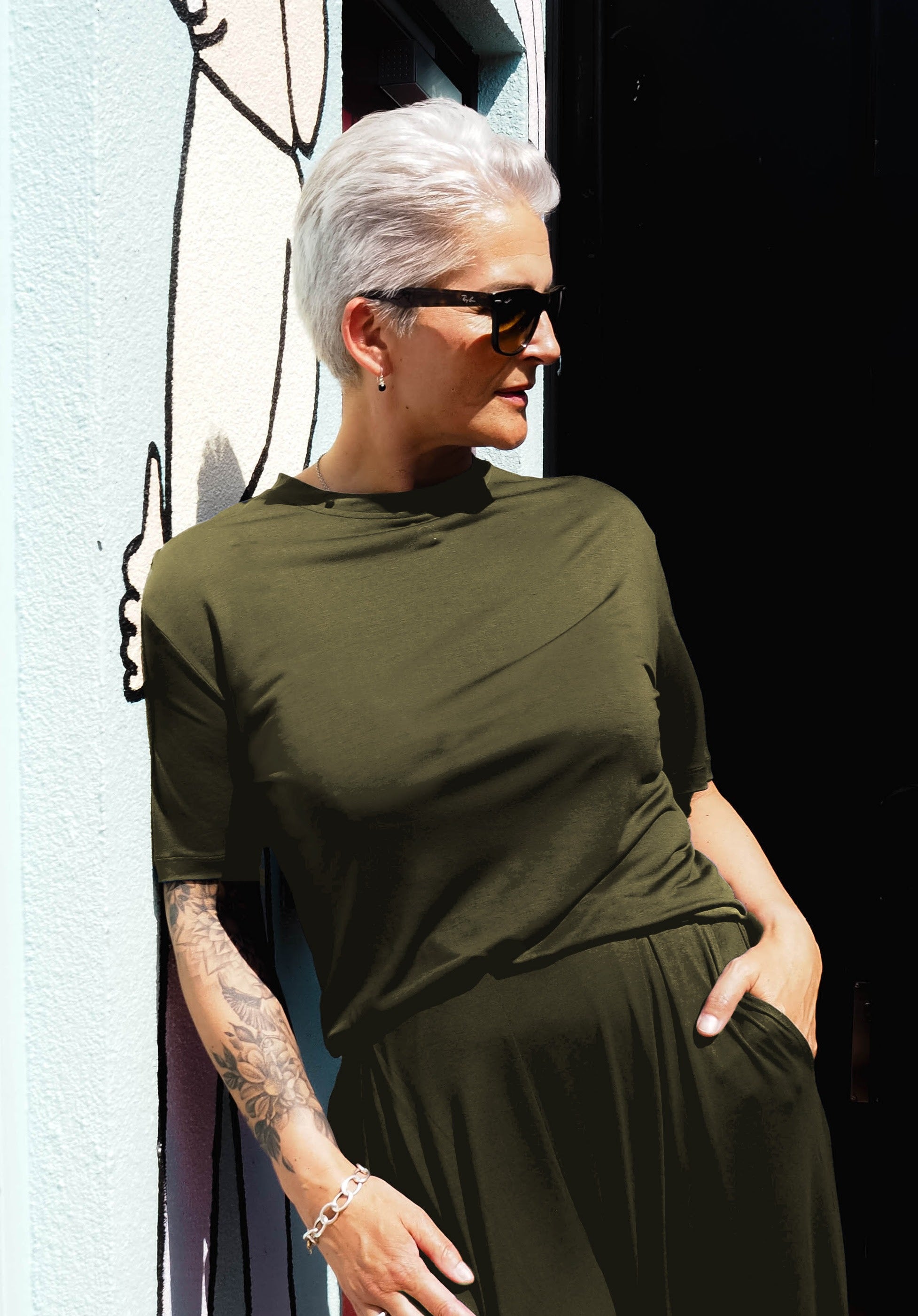 A woman with short, platinum blonde hair stands against a colorful wall, wearing AWAN flow t-shirt and flow pants in olive green. She sports dark sunglasses and a tattooed arm, giving off a cool and modern vibe.