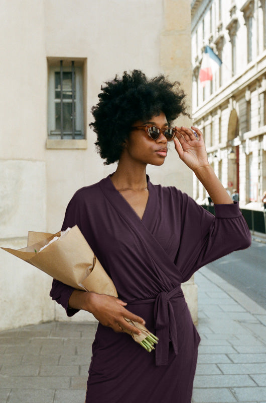 A woman wearing the AWAN Wrap Dress in Deep Plum, holding a bouquet of flowers and adjusting her sunglasses. She is walking down a stylish city street, embodying elegance and versatility in the chic wrap dress.