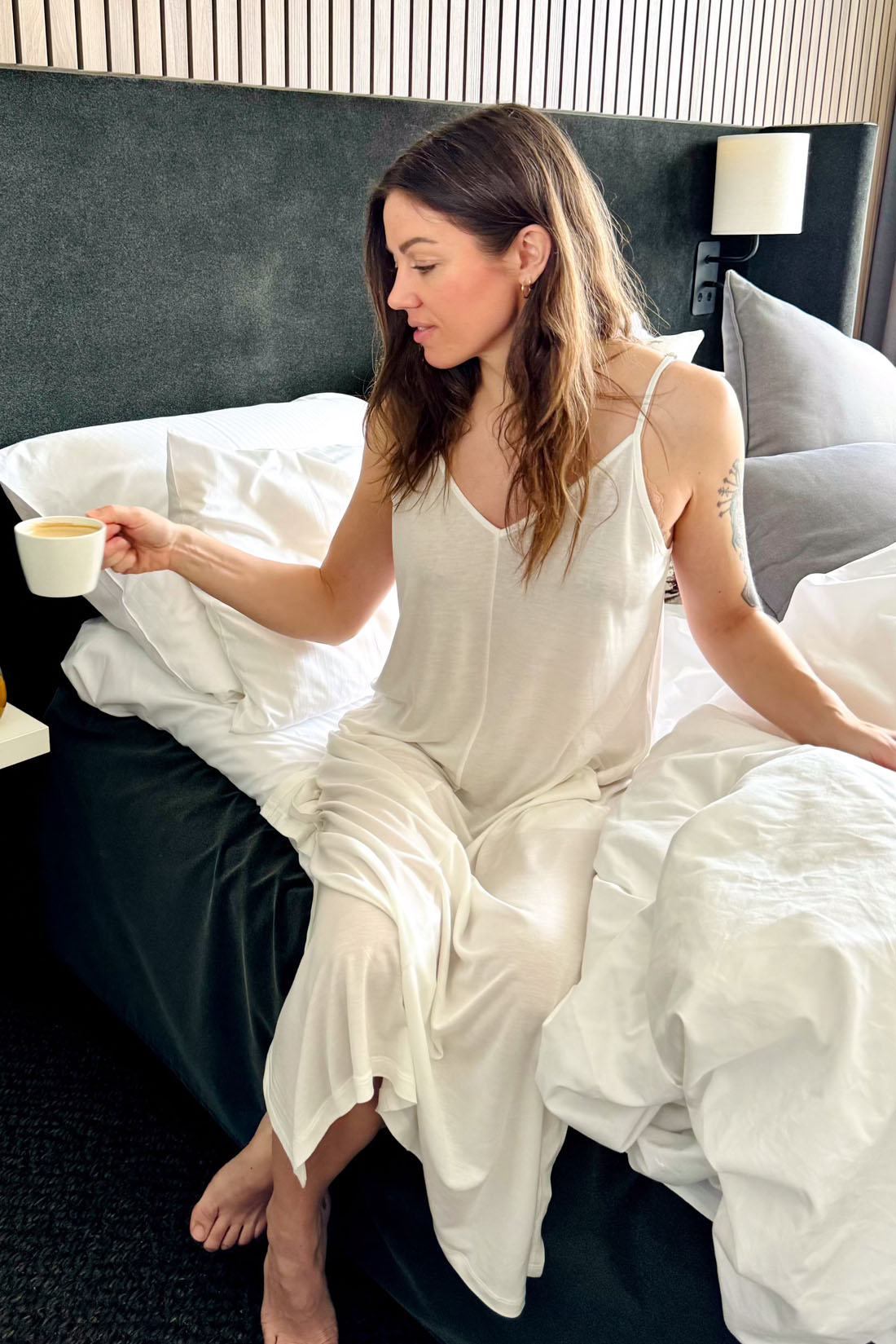 A woman wearing the AWAN Boho Slip Dress in Natural White, sitting on a bed with white linens, holding a cup of coffee, and enjoying a relaxed morning.