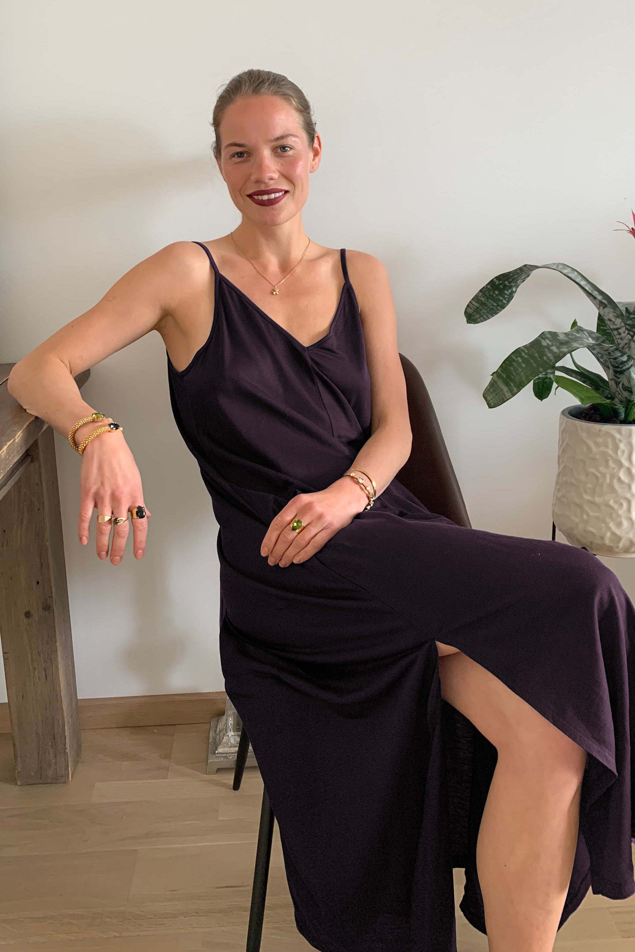 A woman wearing the AWAN Boho Slip Dress in Deep Plum, sitting elegantly on a chair in a modern interior with a potted plant, accessorized with gold jewelry