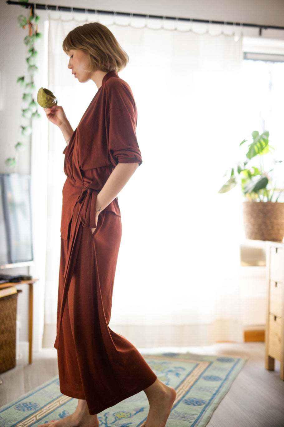 A woman wearing AWAN Flow Pants and a matching wrap top in Deep Rust, standing in a bright room with plants, holding a ceramic cup.