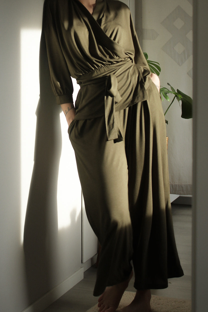 A woman wearing AWAN Flow Pants and a matching wrap top in Olive Green, leaning against a wall with hands in pockets, enjoying natural light.