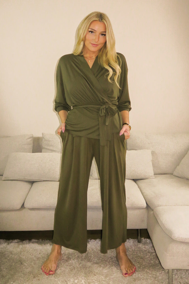 A woman wearing AWAN Flow Pants and a matching wrap top in Olive Green, standing in a cozy living room, smiling and looking comfortable.