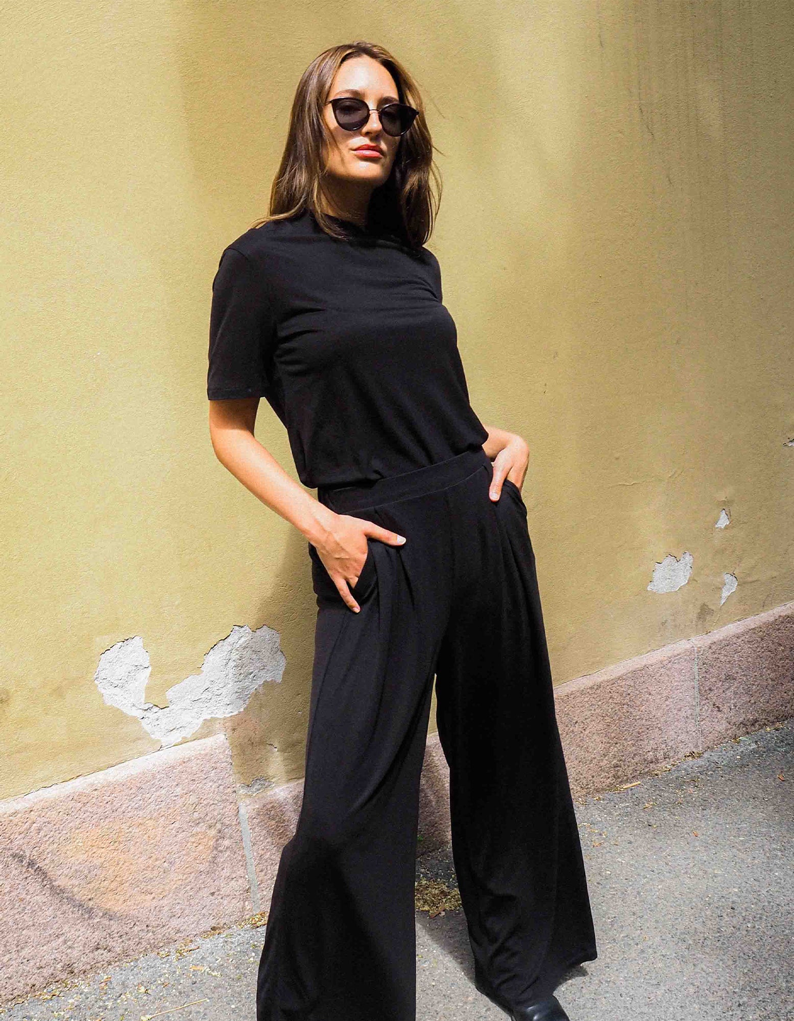 A woman wearing AWAN Flow Pants Long Leg in Warm Black, paired with a matching black top and sunglasses, standing confidently against a textured wall.