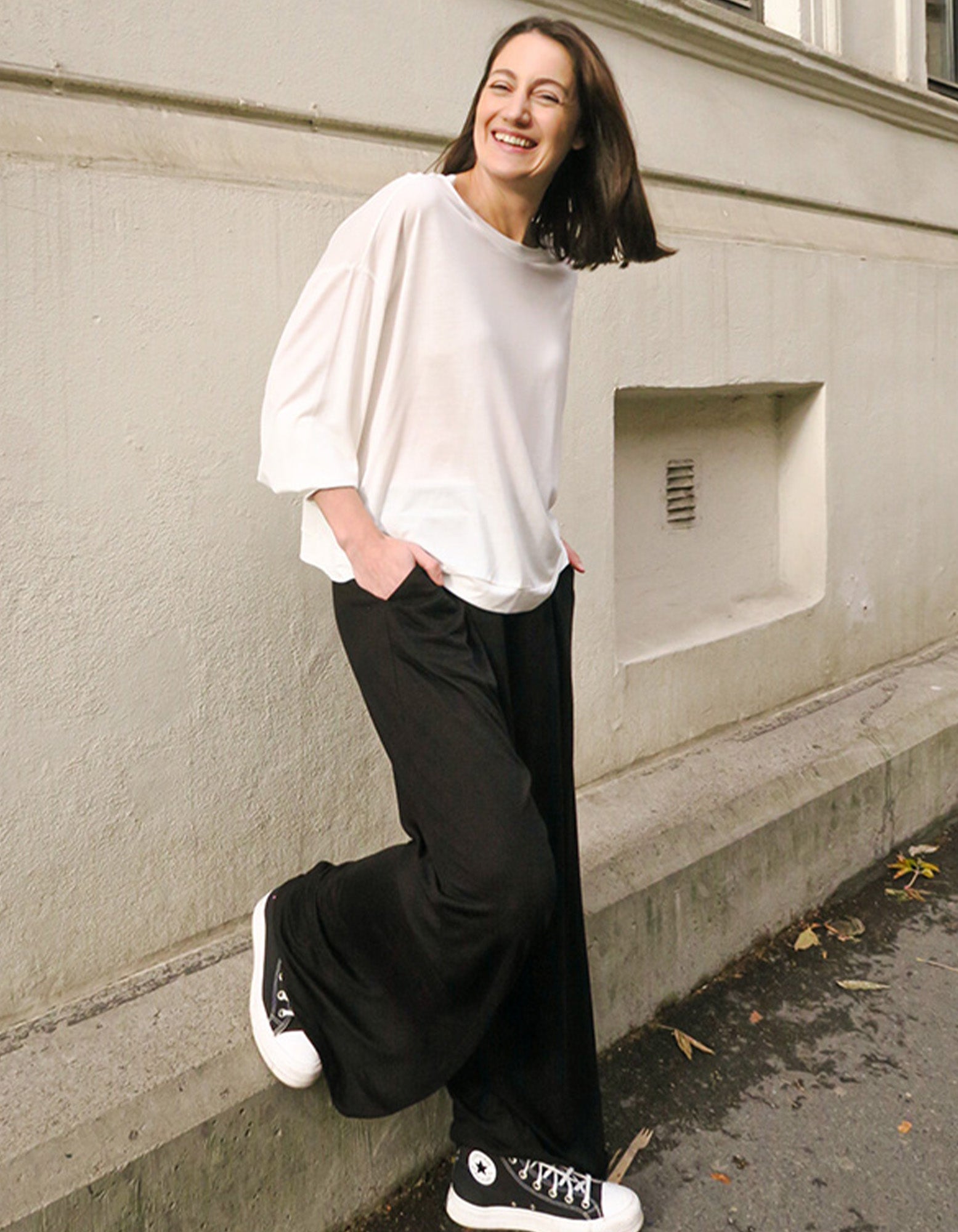 A woman wearing AWAN Flow Pants Long Leg in Warm Black, paired with a white top and black sneakers, leaning against a wall and smiling brightly.