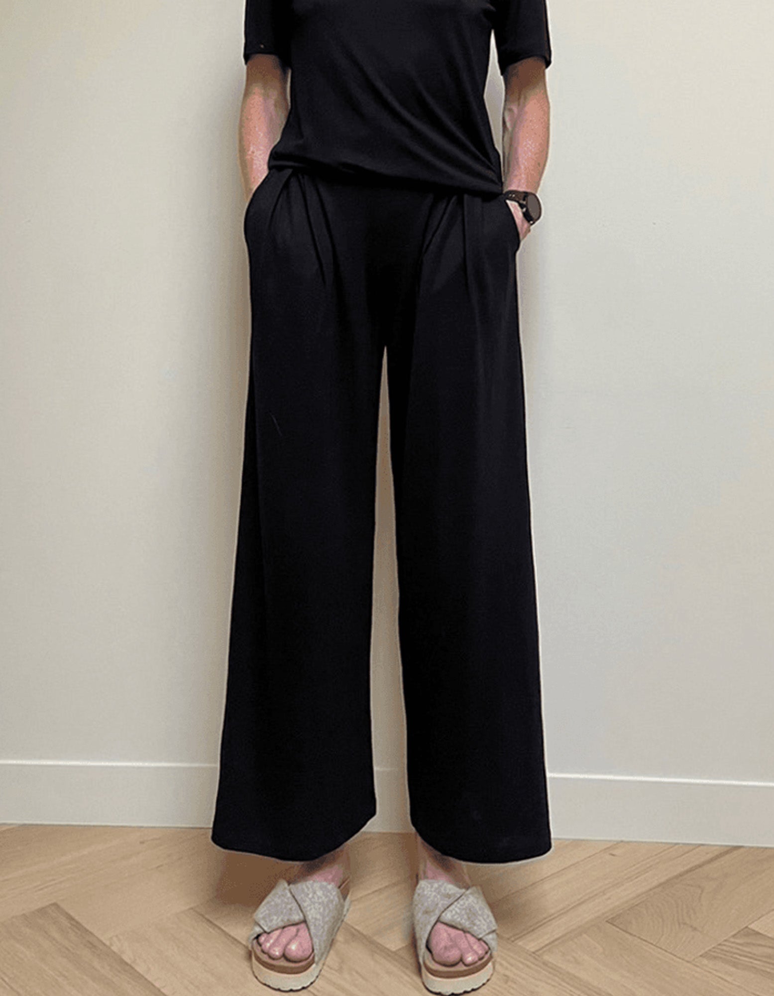 A person wearing AWAN Flow Pants Long Leg in Warm Black, paired with a matching black top and beige sandals, standing against a neutral background.