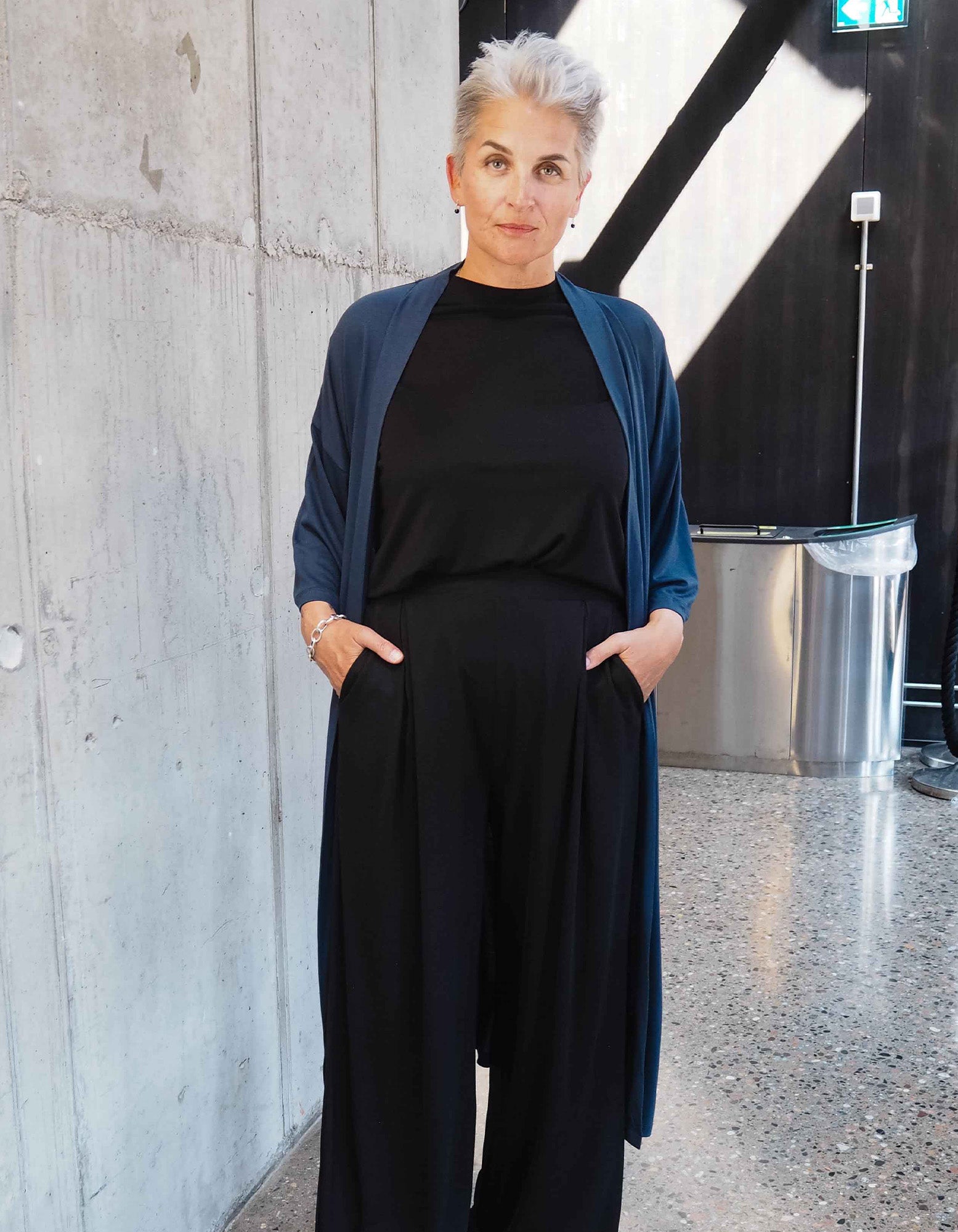 A woman wearing AWAN Flow Pants Long Leg in Warm Black, paired with a black top and a blue cardigan, standing confidently in a modern indoor setting with a concrete wall.