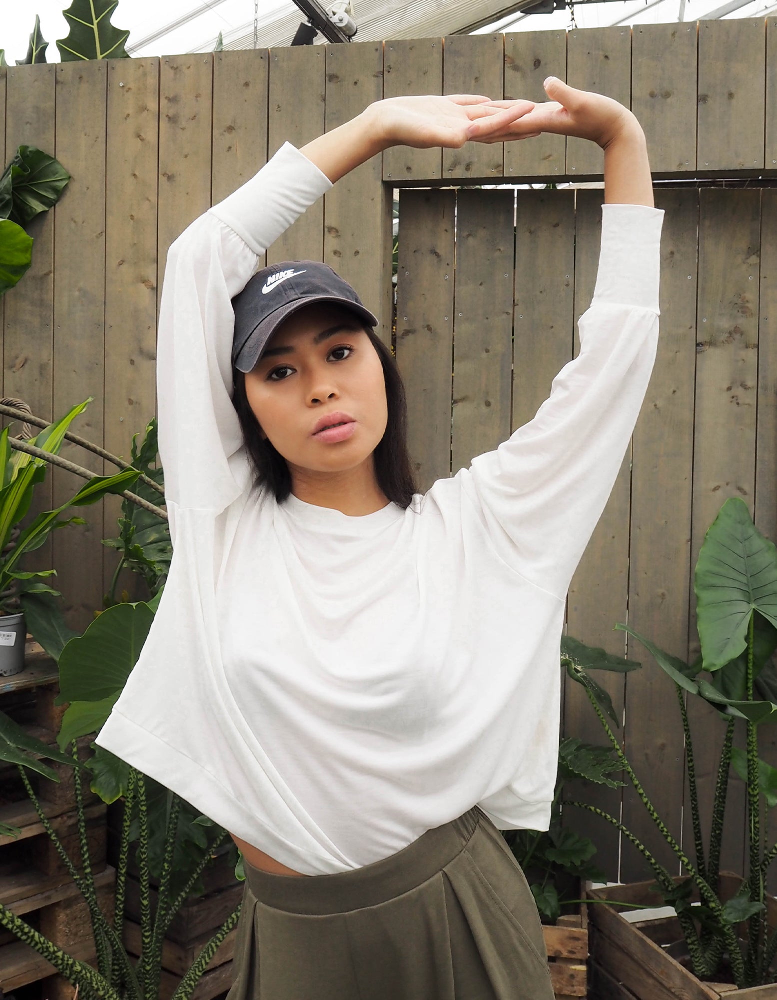 An woman wearing the AWAN Flow Top in natural white, stretching her arms upwards in a garden setting, wearing a black Nike cap and green pants.