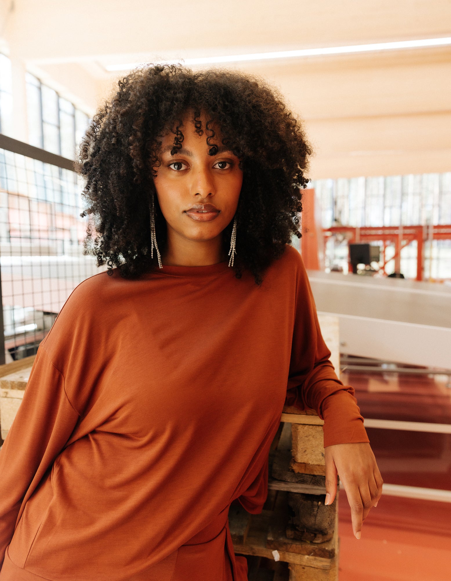 A person dressed in the AWAN Flow Top in deep rust, leaning on a wooden structure indoors, looking directly at the camera with natural light highlighting her features.