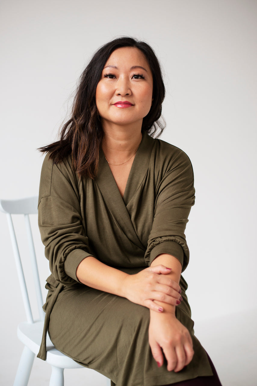 Founder Anette Miwa Dimmen wearing a wrap cardigan and flow pants in olive green, seated on a white chair with a confident smile.