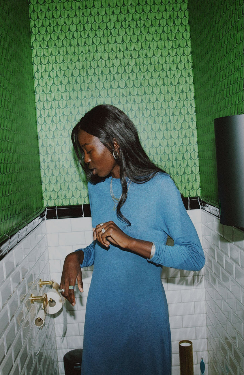 Model wearing AWAN sweater dress in stormy blue, standing in a stylish bathroom with green and white tiled walls.