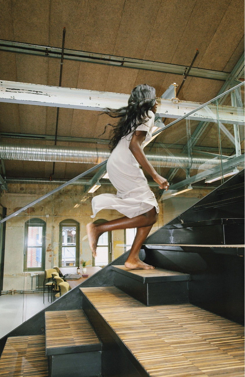 Model wearing AWAN T dress in natural white, running up a modern staircase in an industrial-style building.