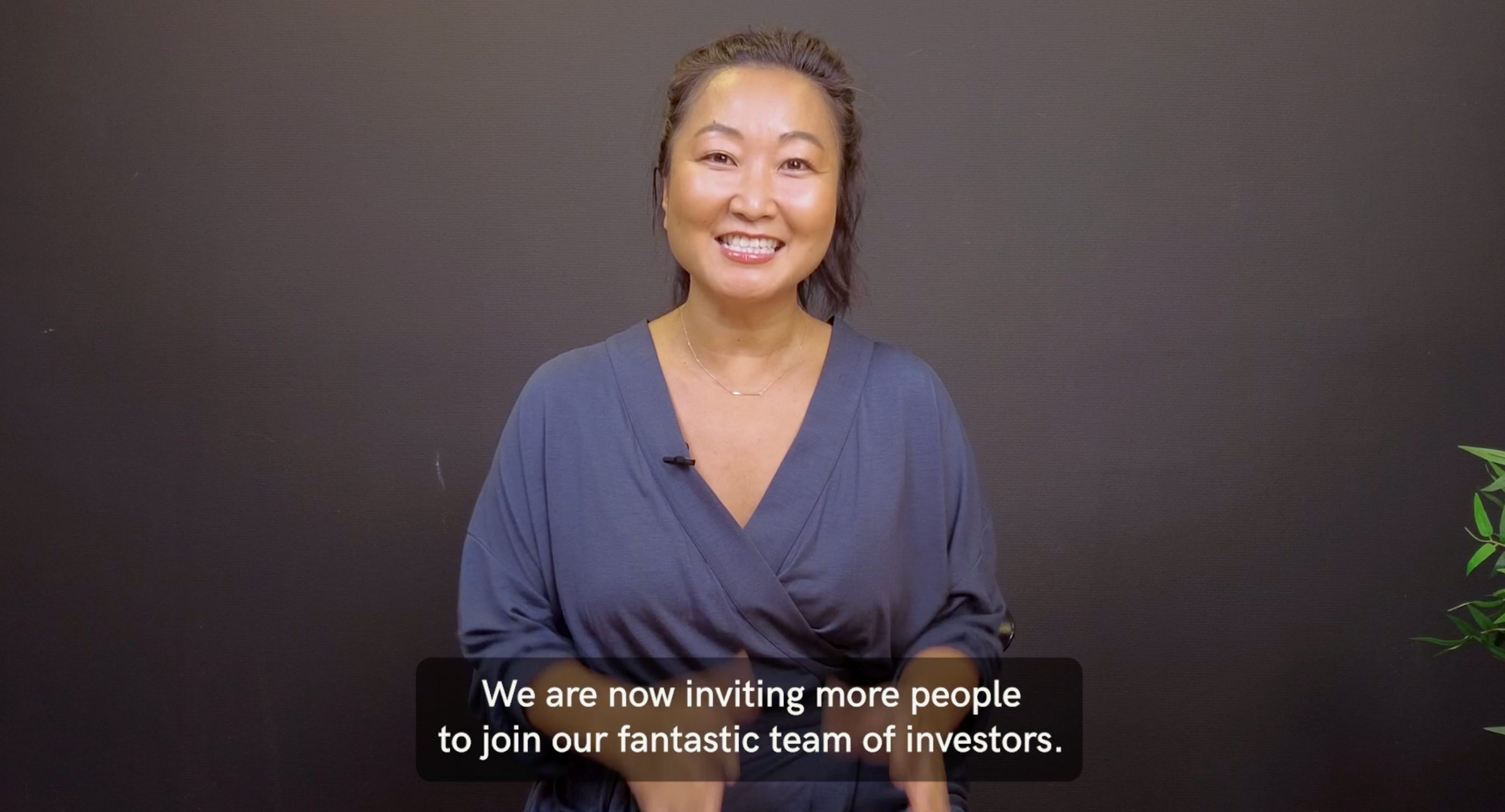 Screenshot from a video featuring Anette Miwa Dimmen wearing a wrap dress in stormy blue, smiling and inviting more people to join the team of investors. Text overlay reads: 'We are now inviting more people to join our fantastic team of investors.