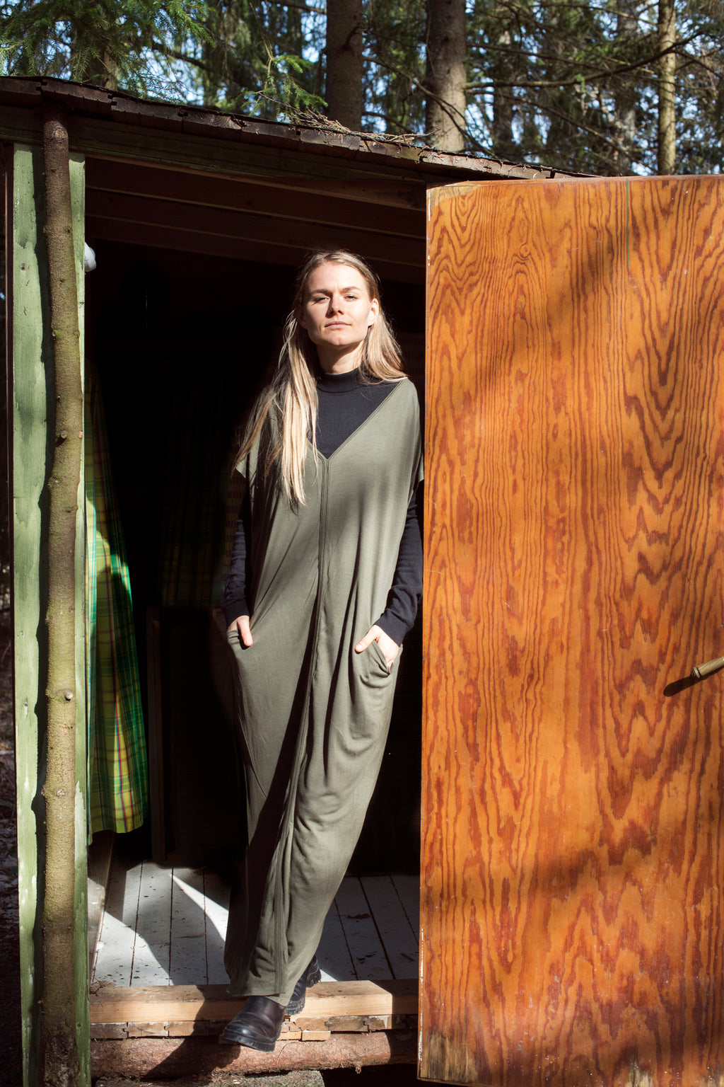 Model standing in front of a rustic wooden door, wearing the AWAN Kaftan Dress in olive green, with hands in pockets.