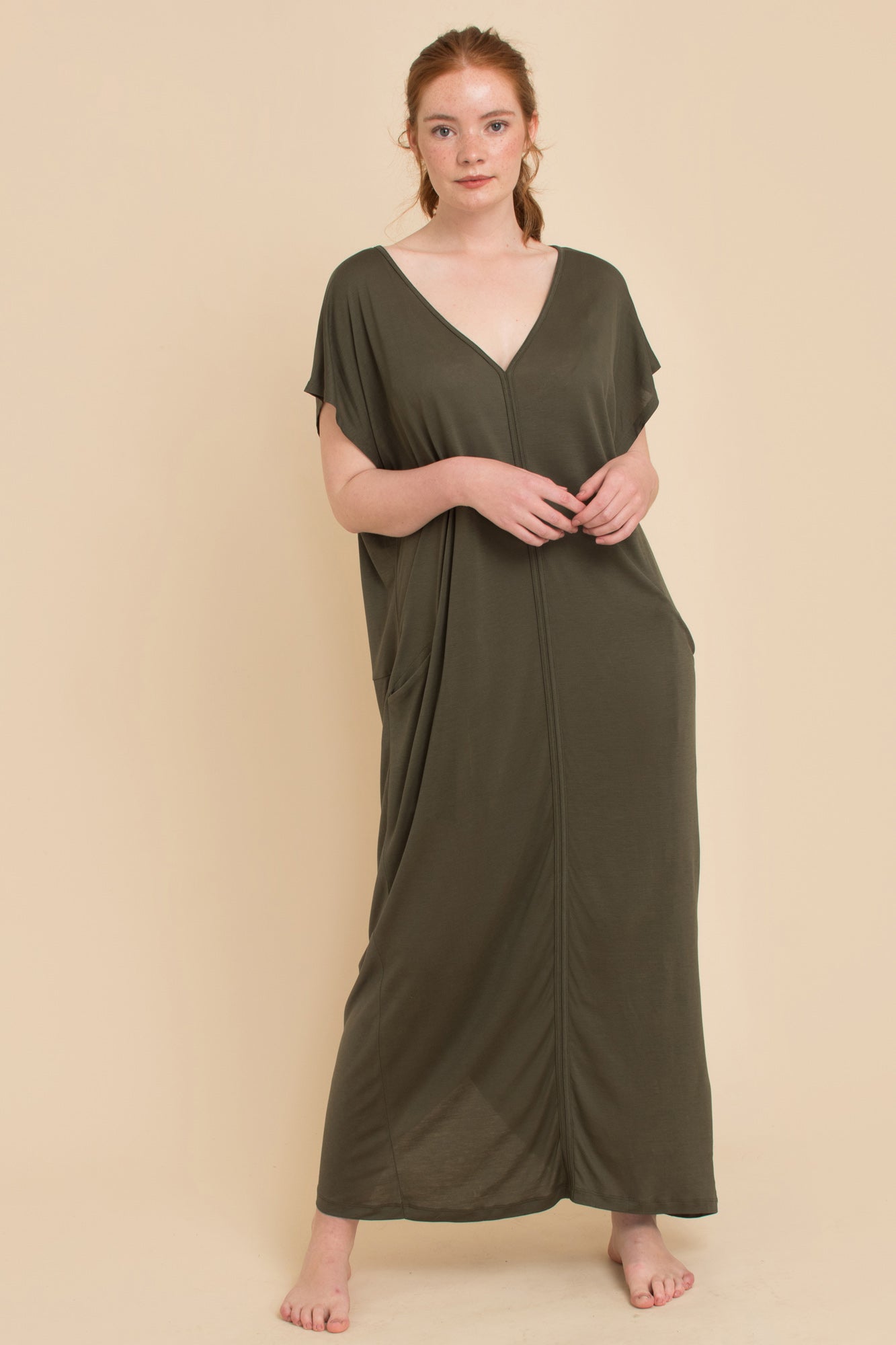 Model posing against a beige background, showcasing the AWAN Kaftan Dress in olive green, with hands in pockets.