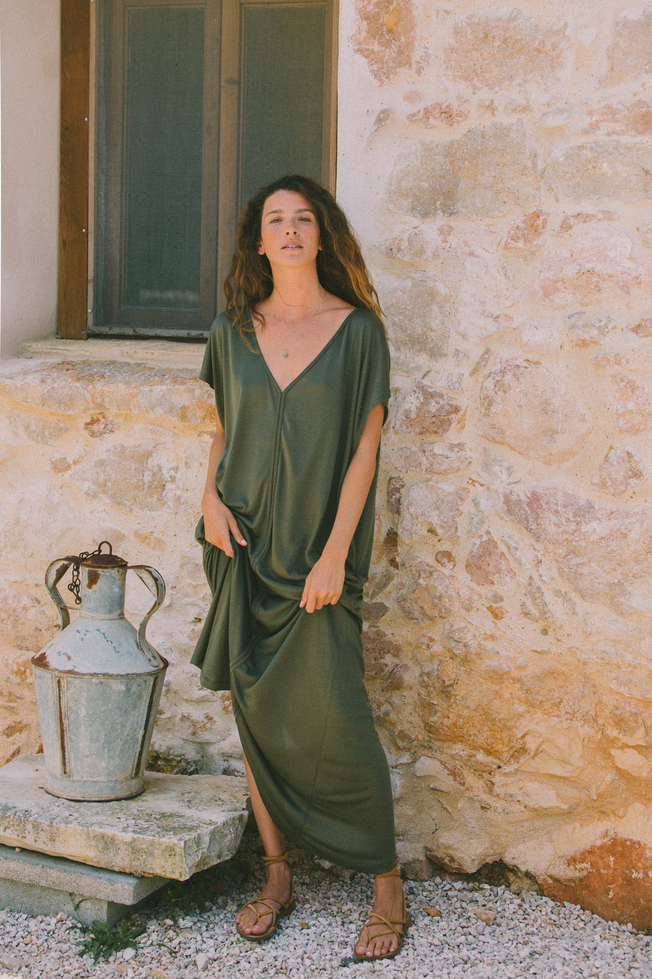 Model standing outdoors against a stone wall, wearing the AWAN Kaftan Dress in olive green, with hands in pockets.