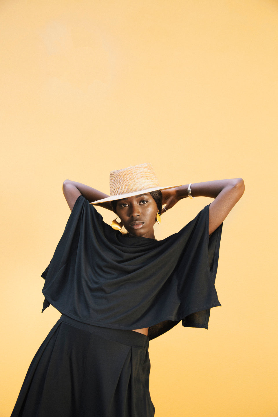 Model striking a pose with arms behind her head, wearing the AWAN Kaftan Top in warm black paired with matching bottoms and a straw hat.