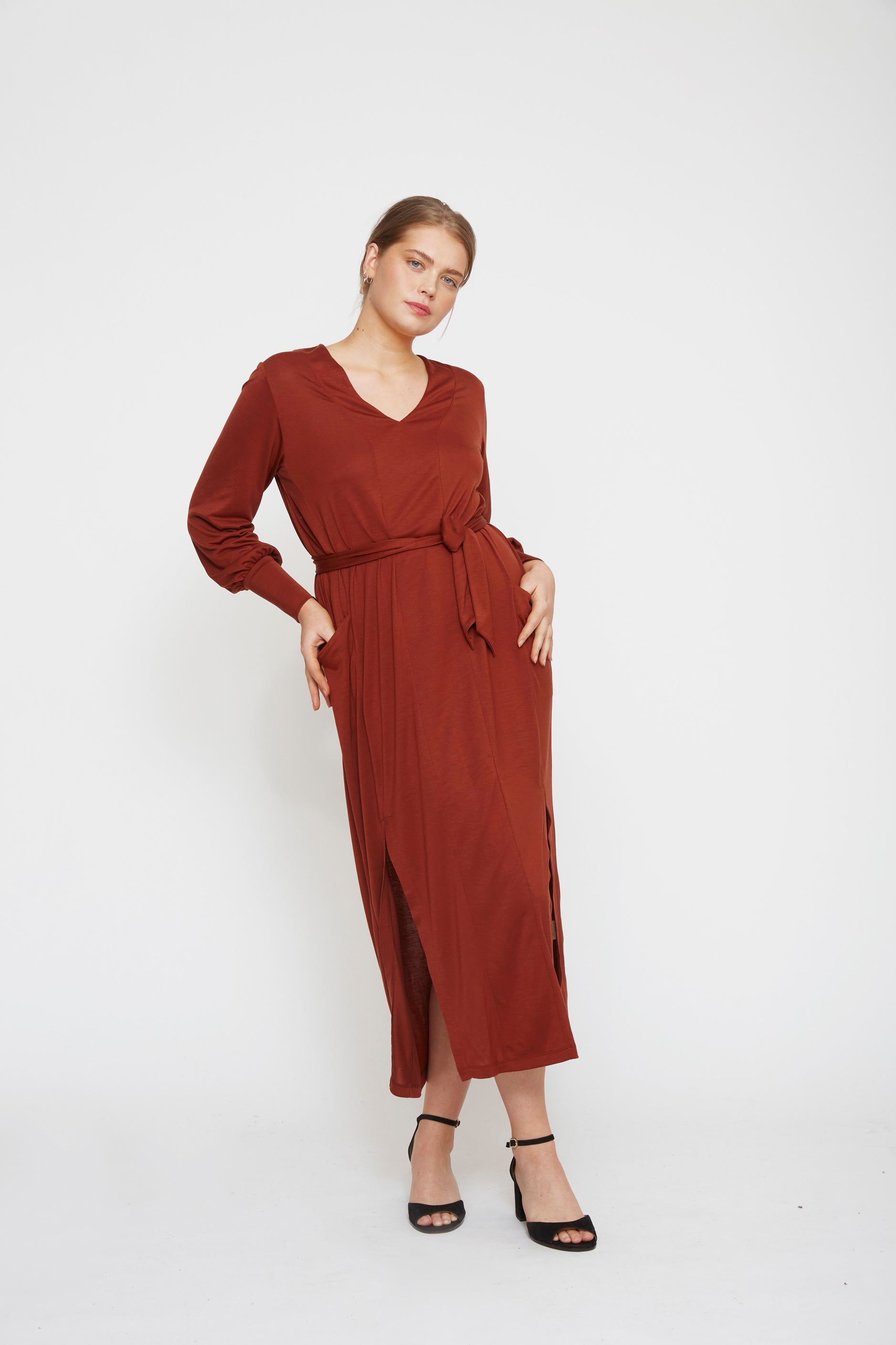 Woman posing in an AWAN long sleeve kaftan dress in deep rust, standing against a white studio backdrop.