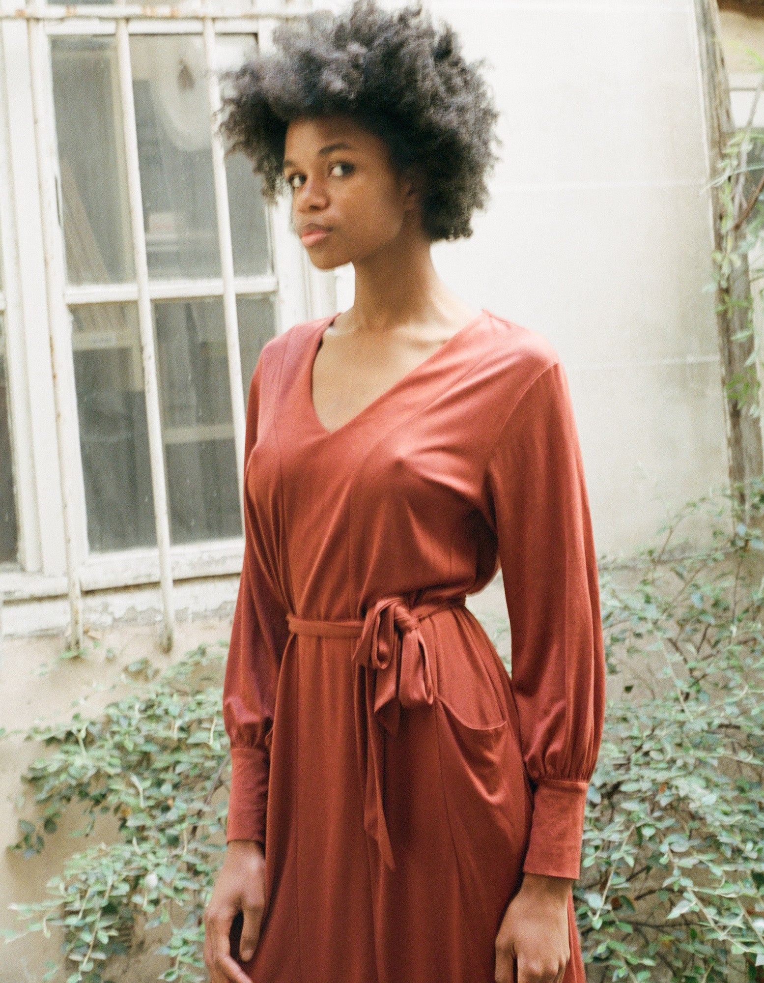 Woman modeling an AWAN long sleeve kaftan dress in deep rust, standing in front of a window with an urban outdoor background.