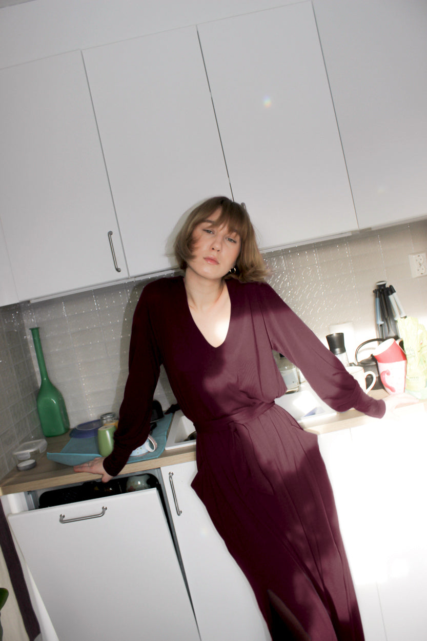 Woman in a kitchen setting wearing an AWAN long sleeve kaftan dress in lush burgundy, leaning against the counter with a casual pose.