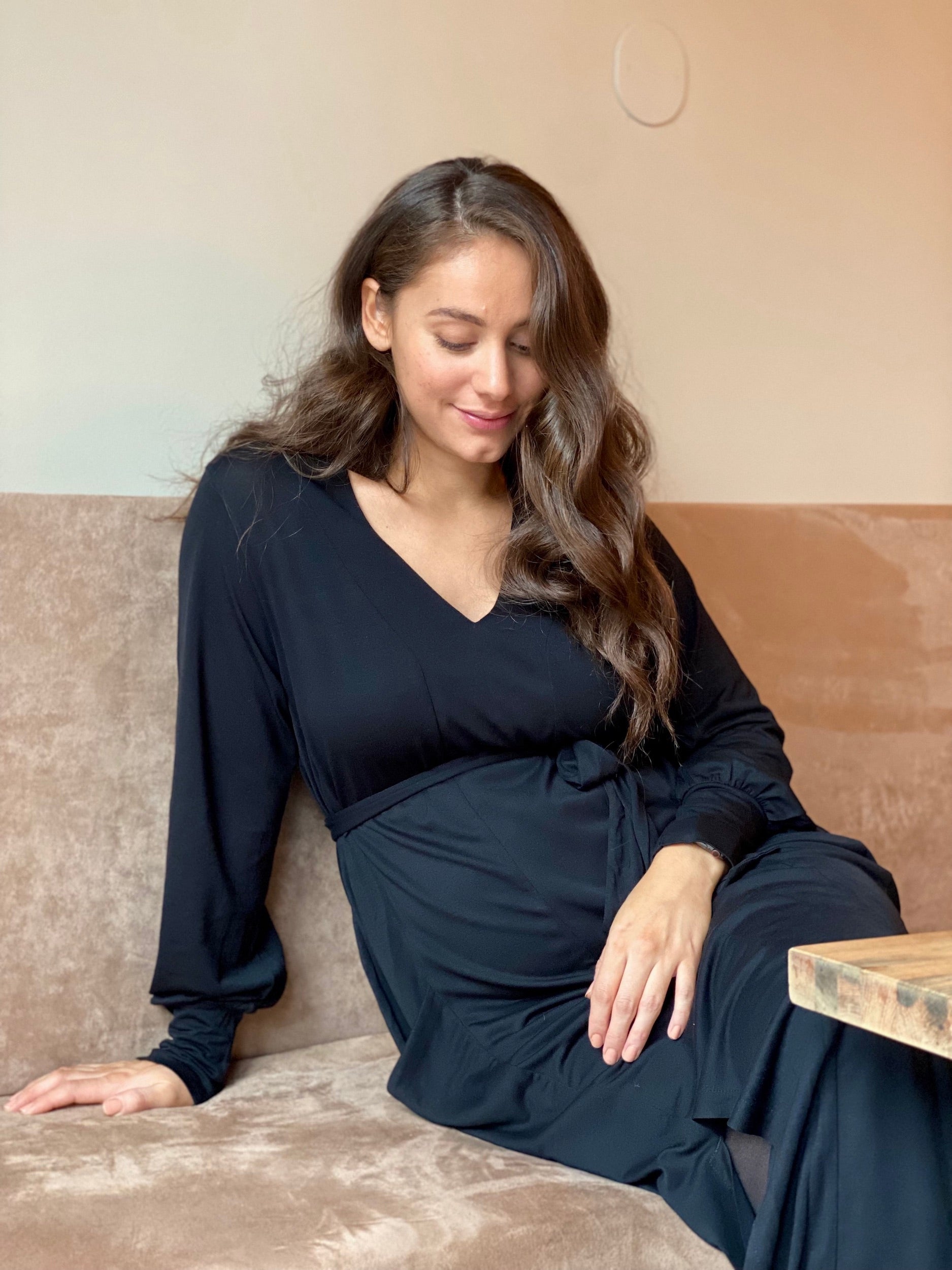 A pregnant woman sits comfortably on a beige couch, wearing an AWAN long sleeve Kaftan dress in warm black. Her hands rest on her baby bump, highlighting the elegance and comfort of the maternity dress.