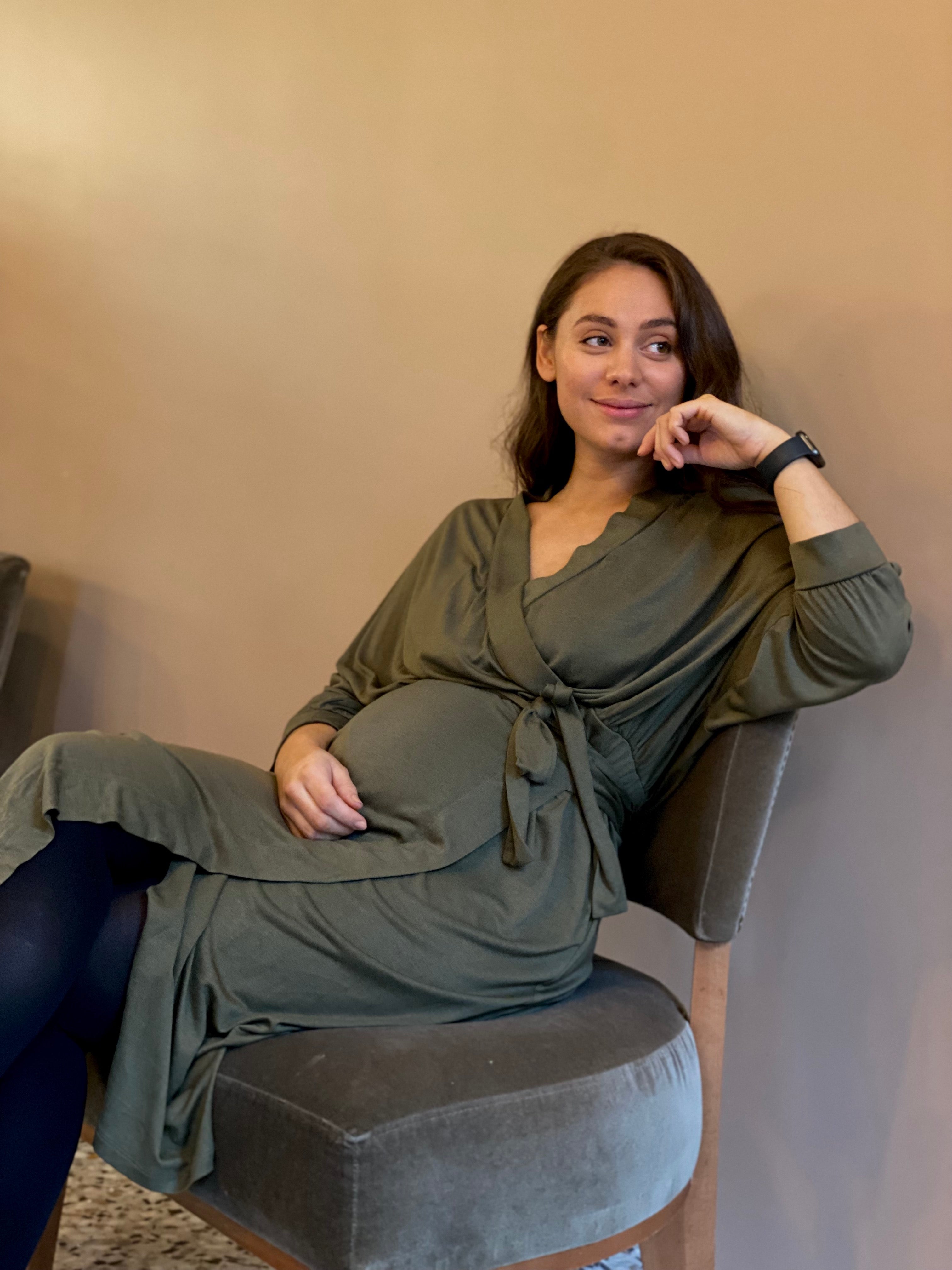 A model is seated on a chair, wearing the AWAN Wrap Dress in Olive Green, with a serene expression and a hand resting on her chin.