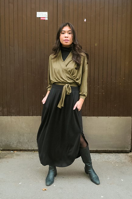 : A woman stands in front of a wooden wall, wearing a warm black midi flow skirt paired with an olive green wrap top, her hands in her skirt pockets.