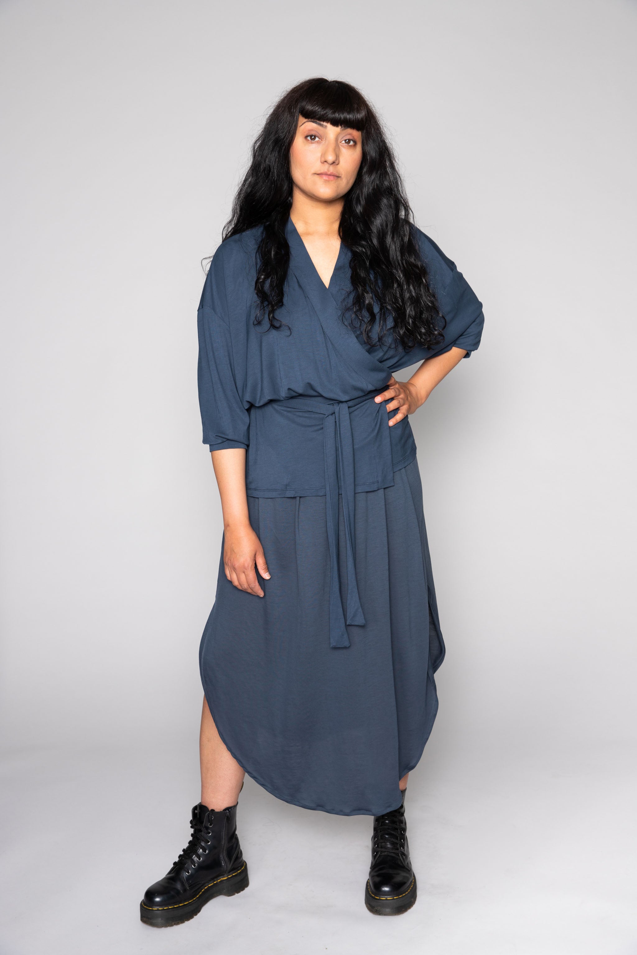 A woman poses in a studio wearing a stormy blue wrap top and matching midi flow skirt, paired with black combat boots.