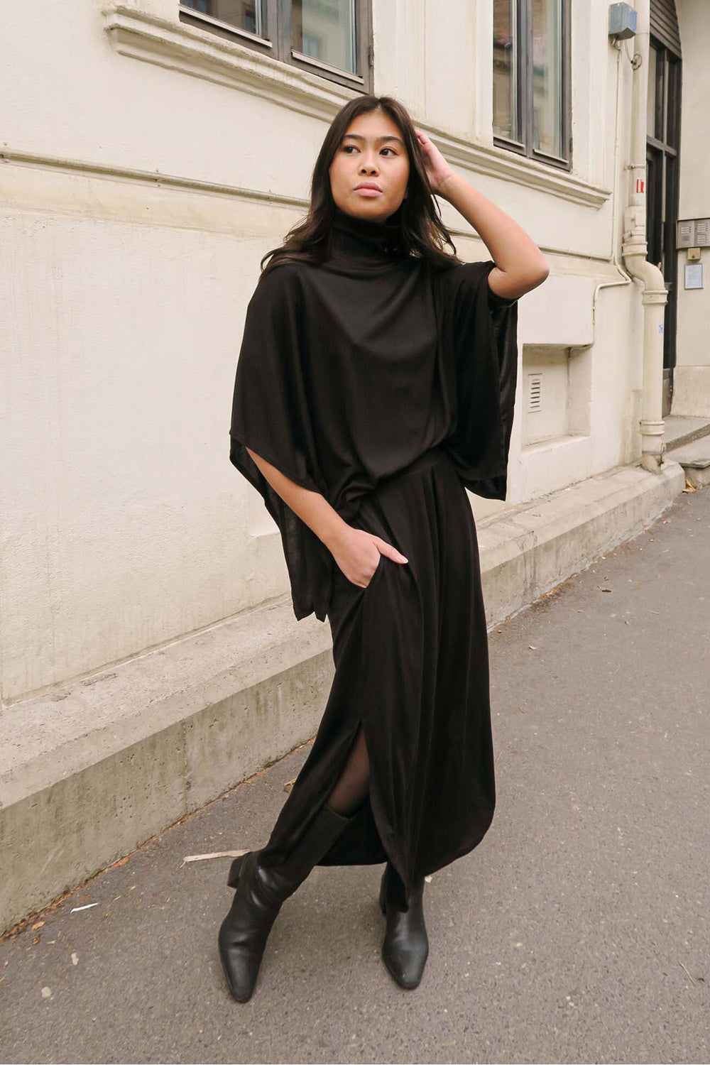 A woman stands outdoors against a beige building, wearing a warm black midi flow skirt paired with a high-neck top, her hand running through her hair.