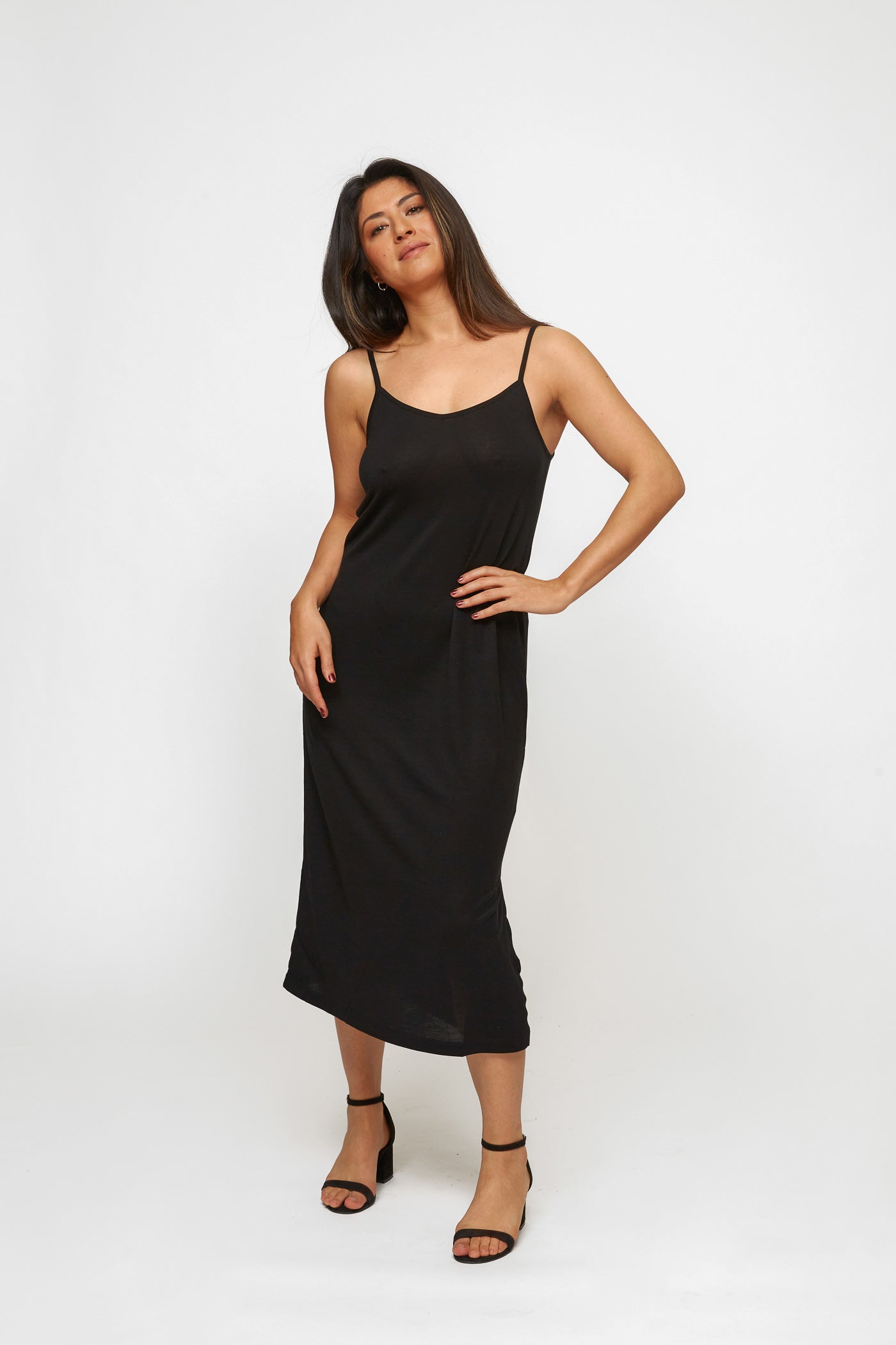 A woman posing confidently in a black midi slip dress, her hand on her hip, with a plain white background highlighting the dress.