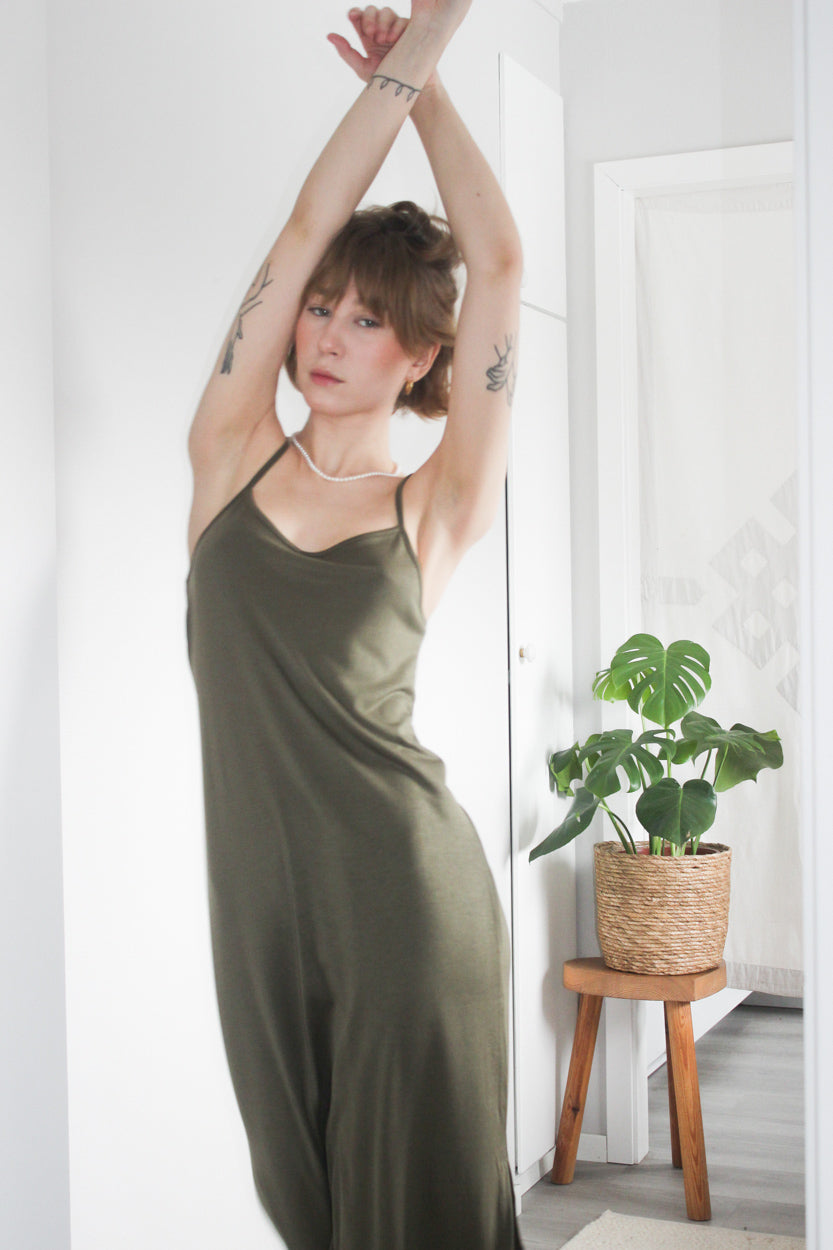 Woman with arms raised, wearing an olive green midi slip dress, standing in a modern, minimalist room with a potted plant on a wooden stool.