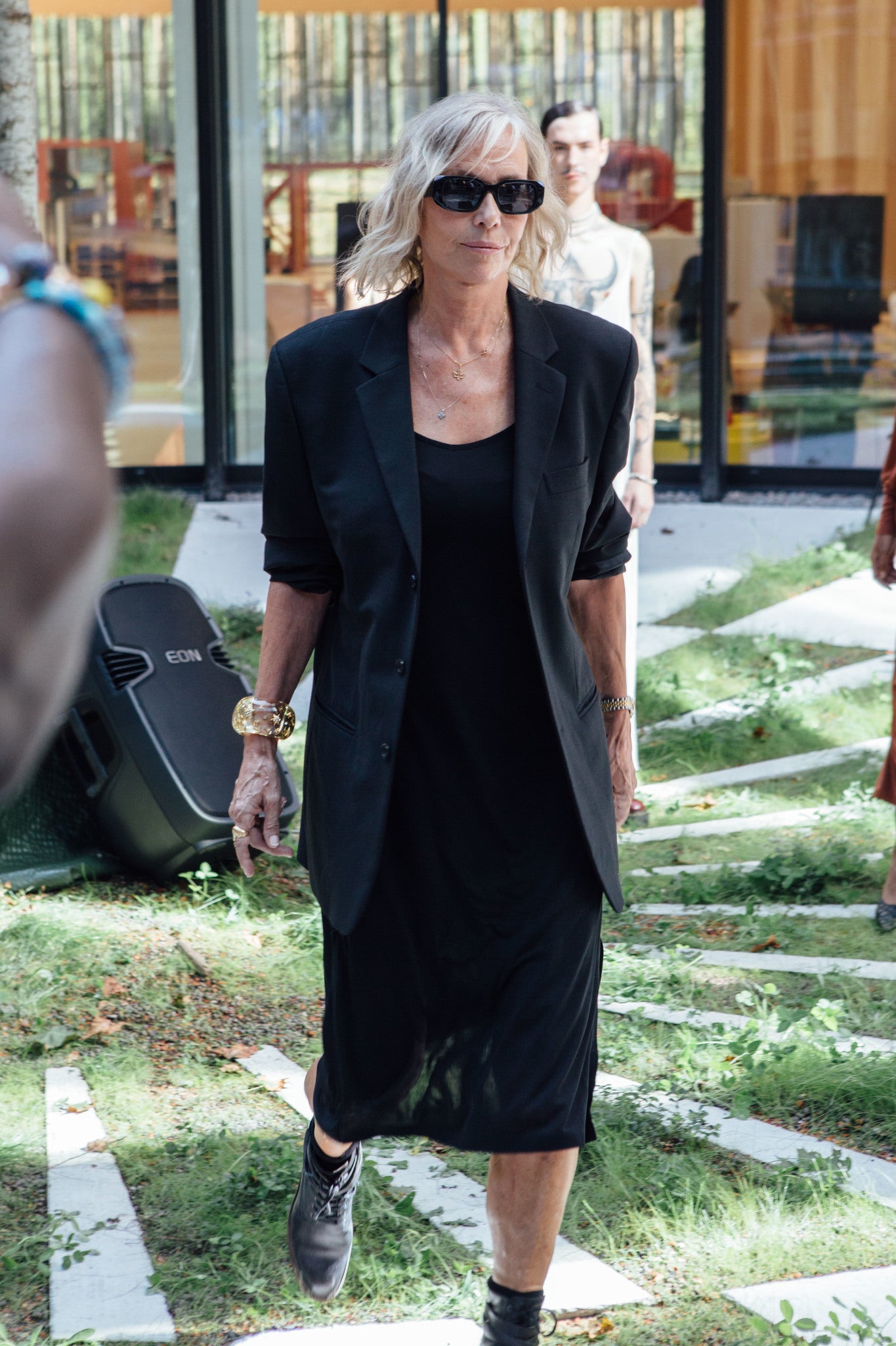 Street style shot of a woman wearing a black midi slip dress paired with a black blazer, sunglasses, and chunky jewelry, walking confidently in an urban setting.