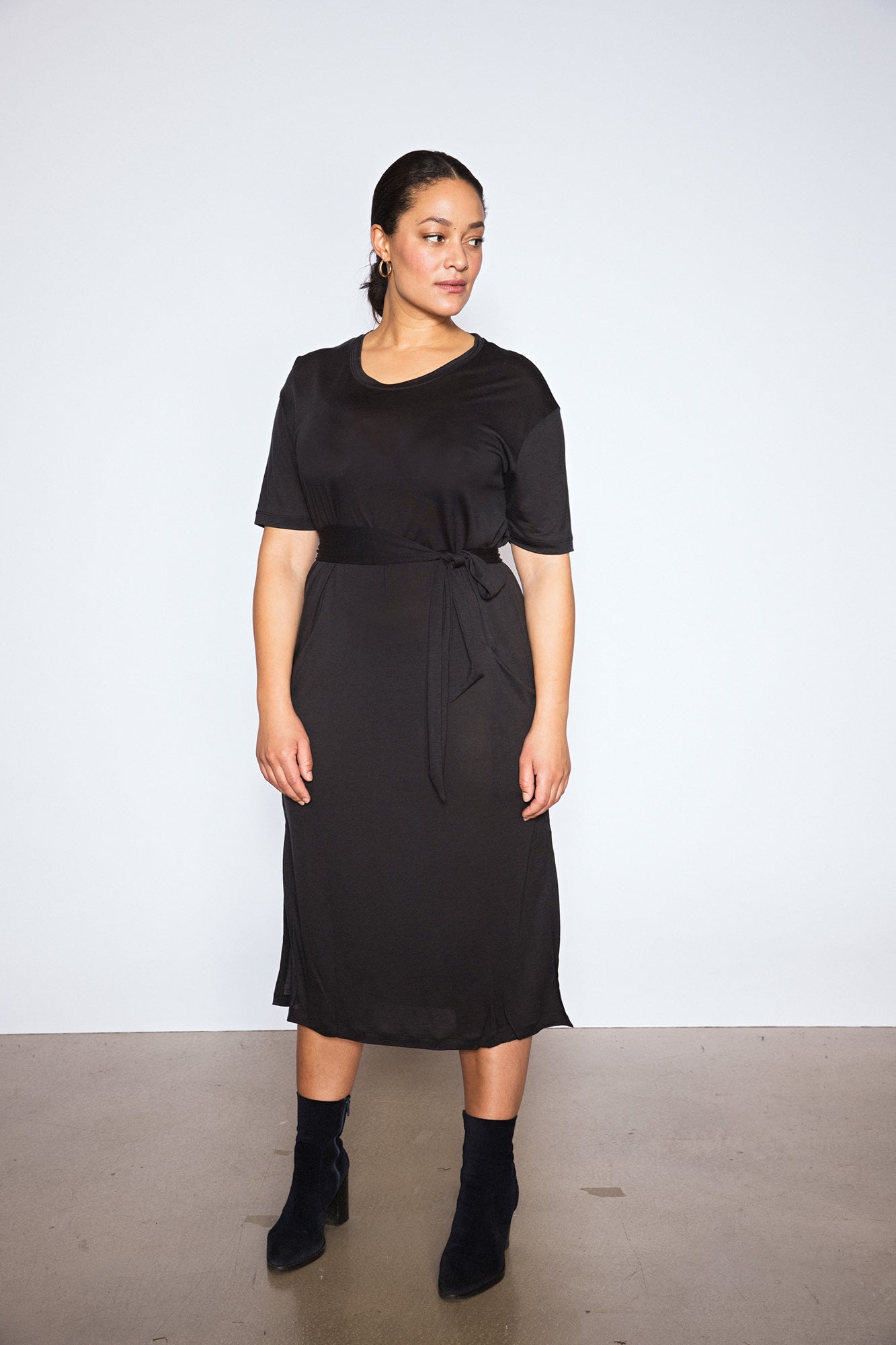 Woman wearing a Midi T dress in warm black. The dress has short sleeves and a tie belt at the waist, paired with black ankle boots. The woman stands against a plain white background.