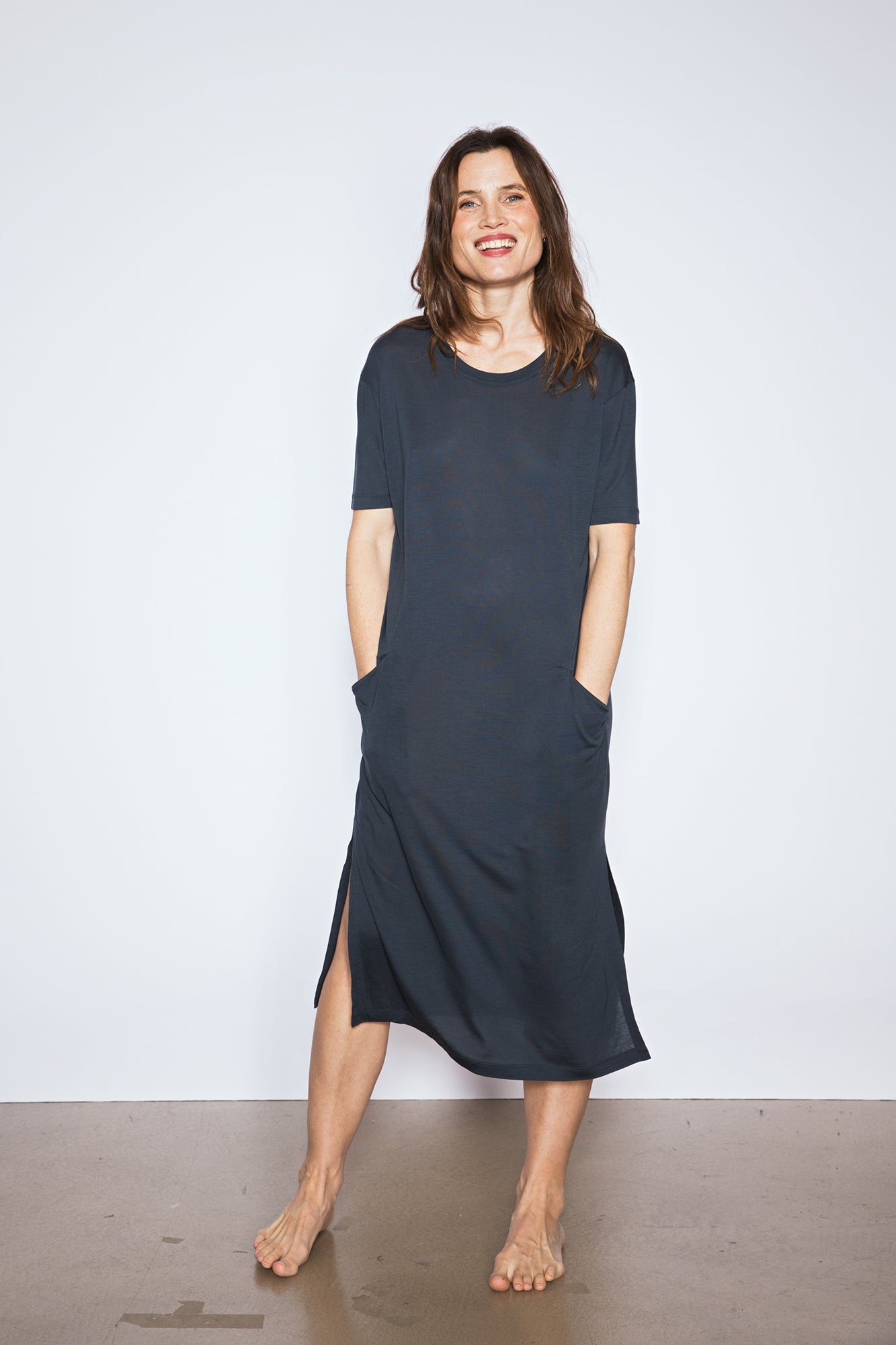 Woman wearing a Midi T dress in stormy blue. The dress features short sleeves and pockets. The woman stands barefoot against a plain white background, smiling with her hands in her pockets.