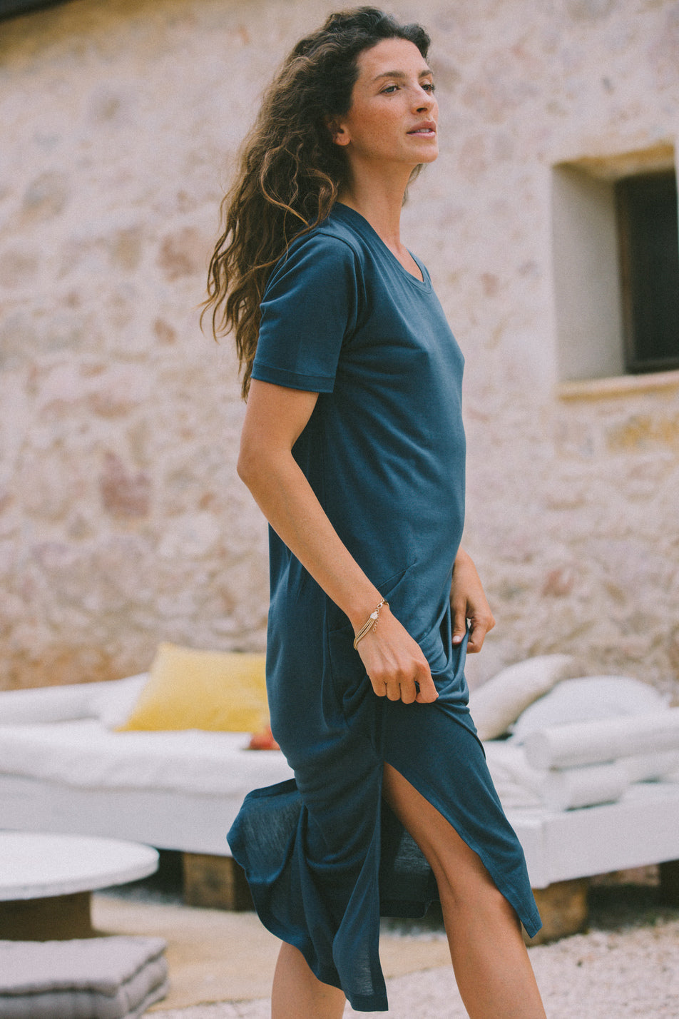 Woman wearing a Midi T dress in stormy blue. The dress features short sleeves and pockets. The woman stands barefoot against a plain white background, smiling with her hands in her pockets.