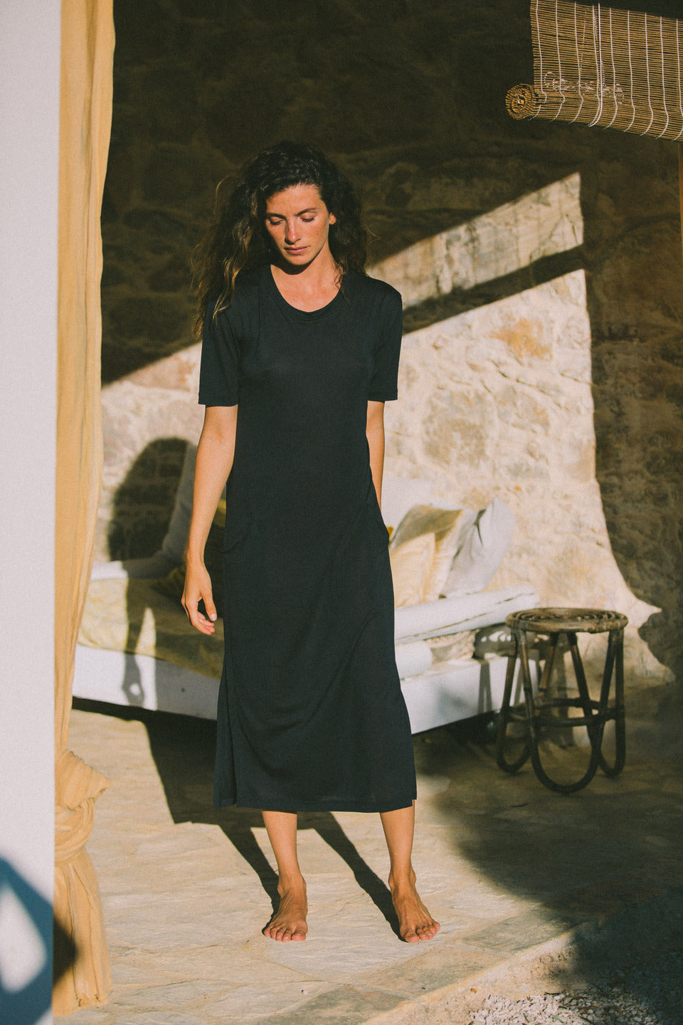 Woman wearing a Midi T dress in stormy blue outdoors. The dress has short sleeves and a side slit, and the woman walks barefoot in a rustic outdoor setting with stone walls.