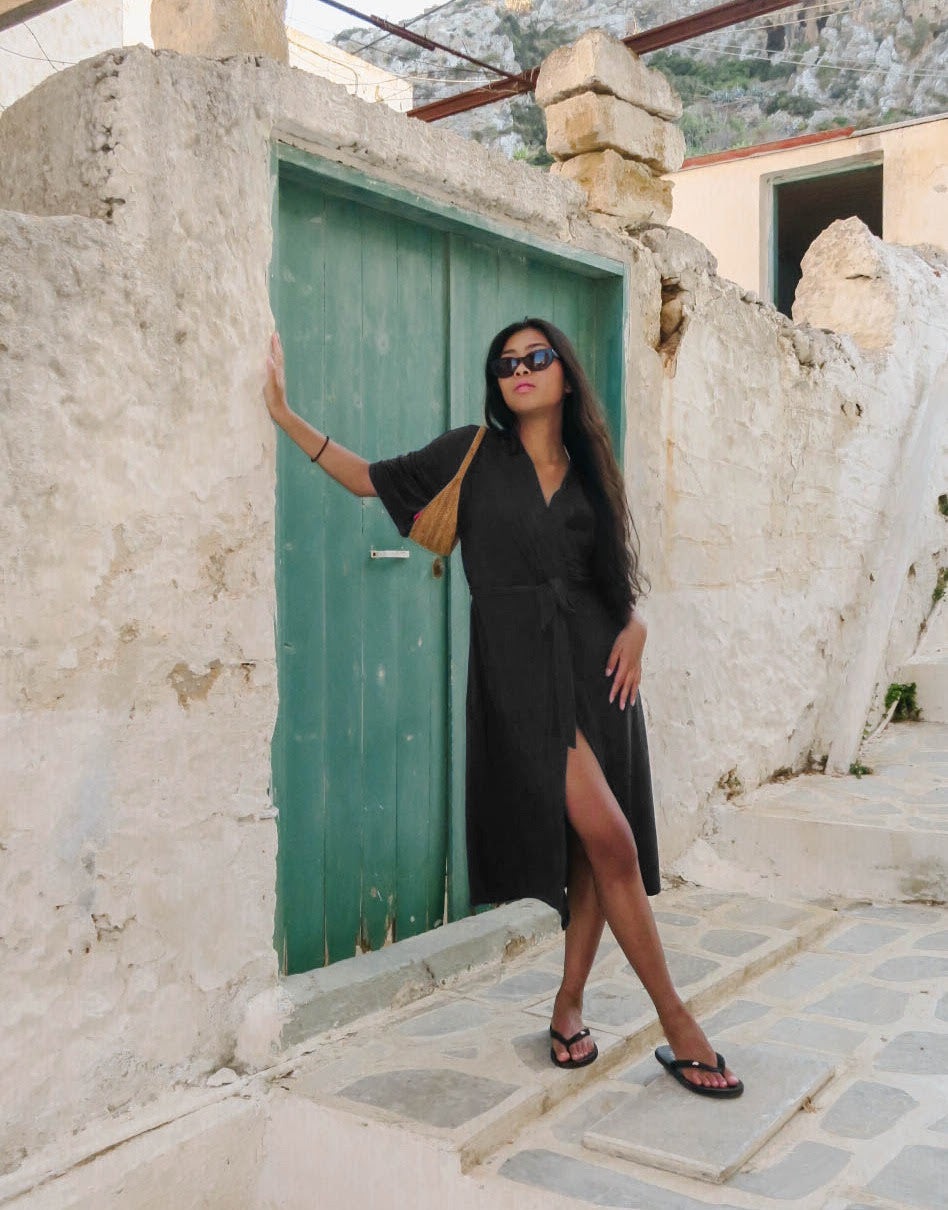  A stylish woman leans against a rustic stone wall with a teal door, wearing AWAN Kimono robe in olive green. She accessorizes with black sunglasses, flip-flops, and a shoulder bag, embodying a casual and effortless style.