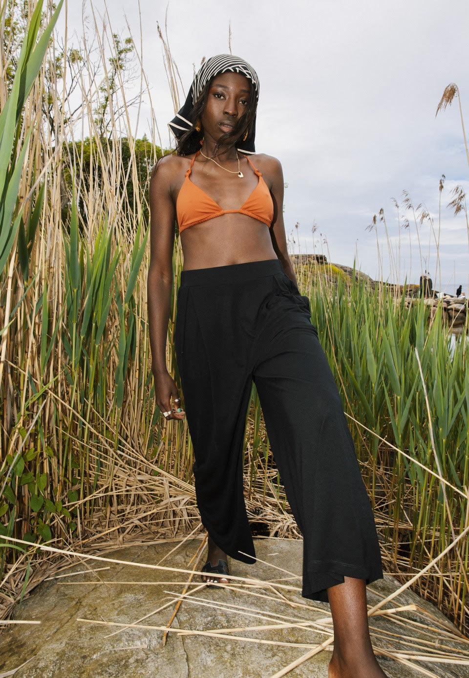 A model stands in a natural setting among tall grasses, wearing AWAN flow pants in warm black. She pairs the pants with an orange bikini top and a patterned headscarf, highlighting a stylish and relaxed beach look.
