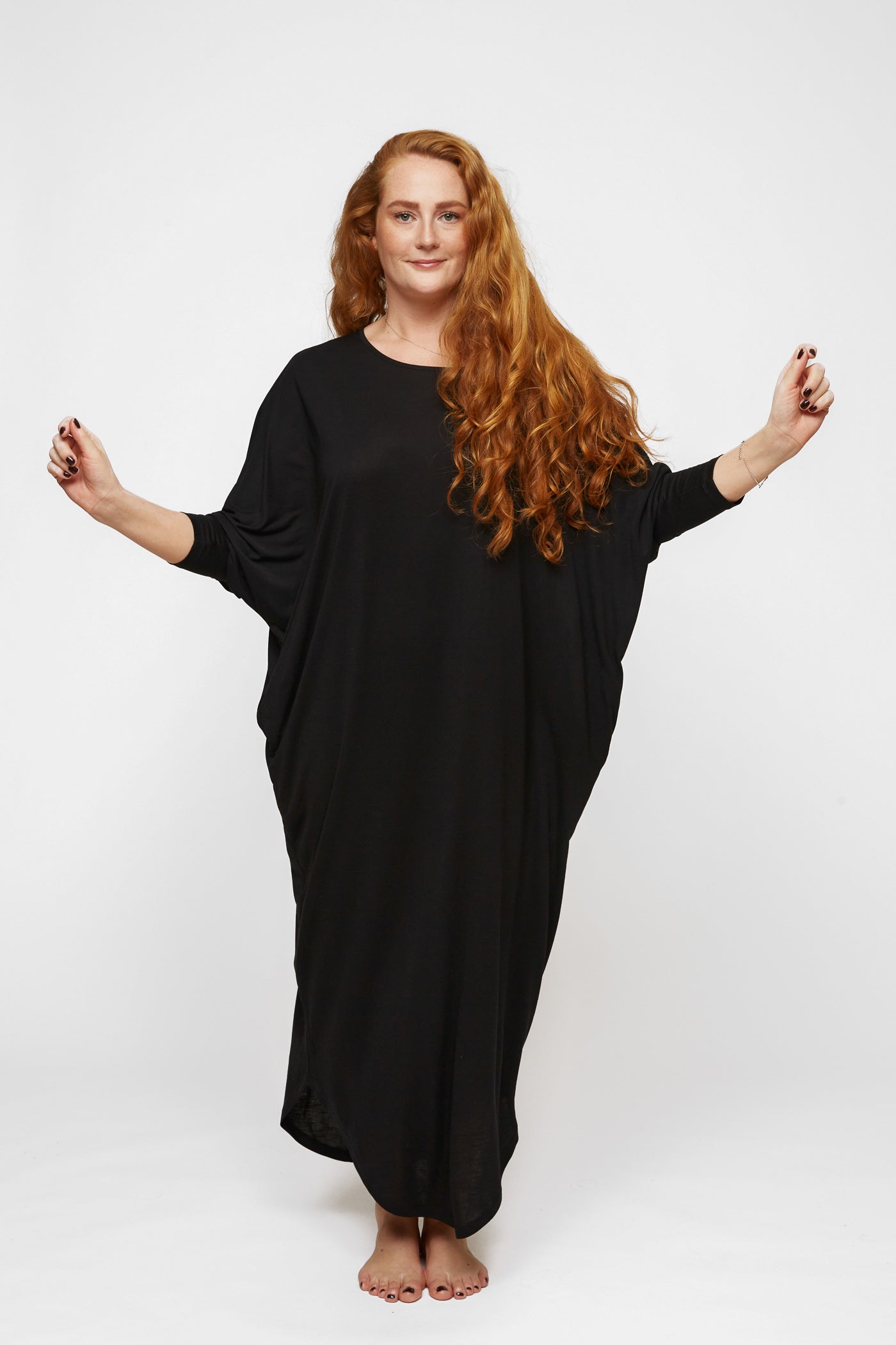 A woman with long red hair stands against a white background, wearing an AWAN Sweater Kaftan dress in warm black. She spreads her arms out and smiles.