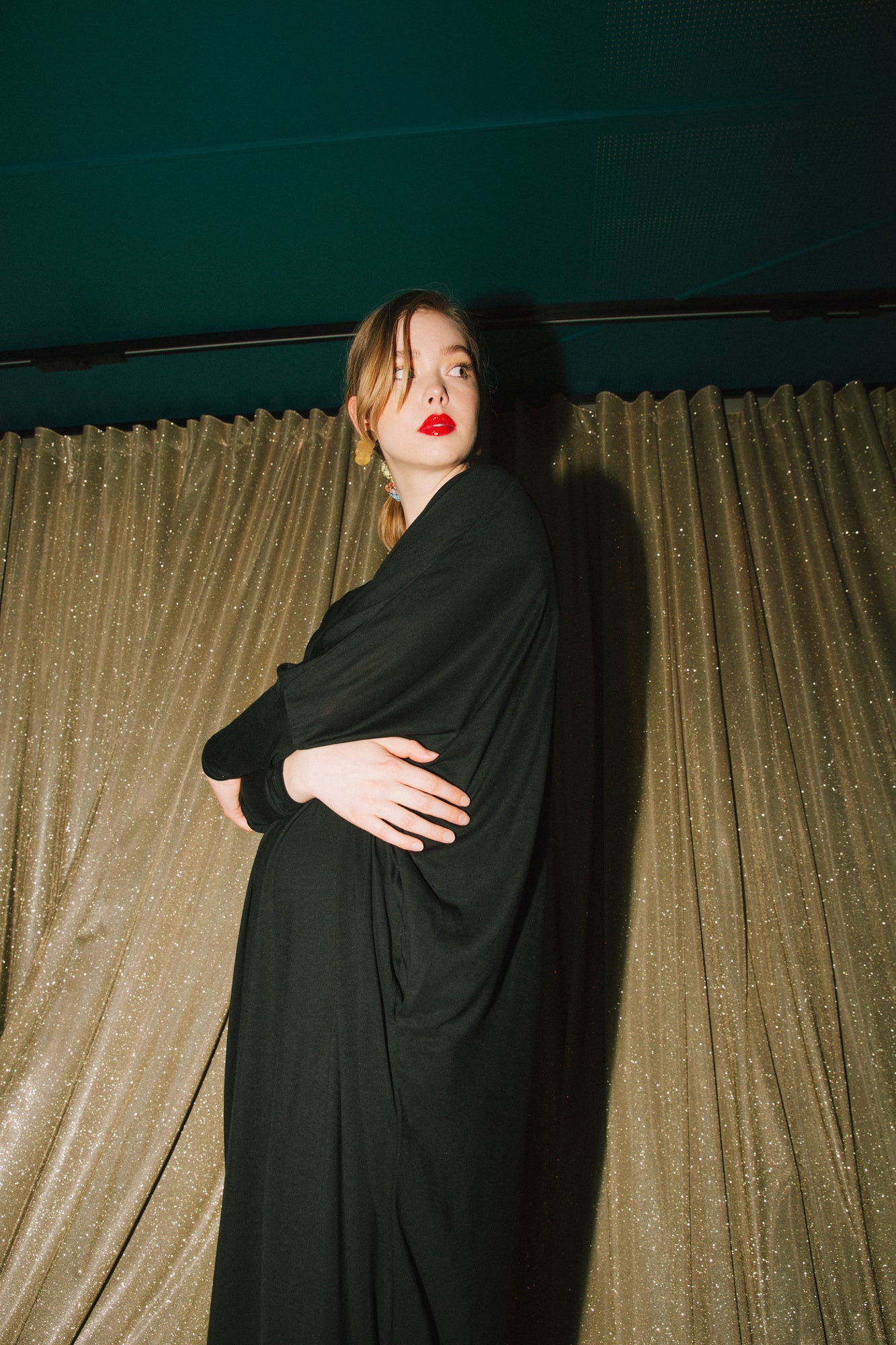 A woman stands against a glittery curtain, wearing an AWAN Sweater Kaftan dress in warm black. She has her arms crossed and is looking to the side with red lipstick and earrings.