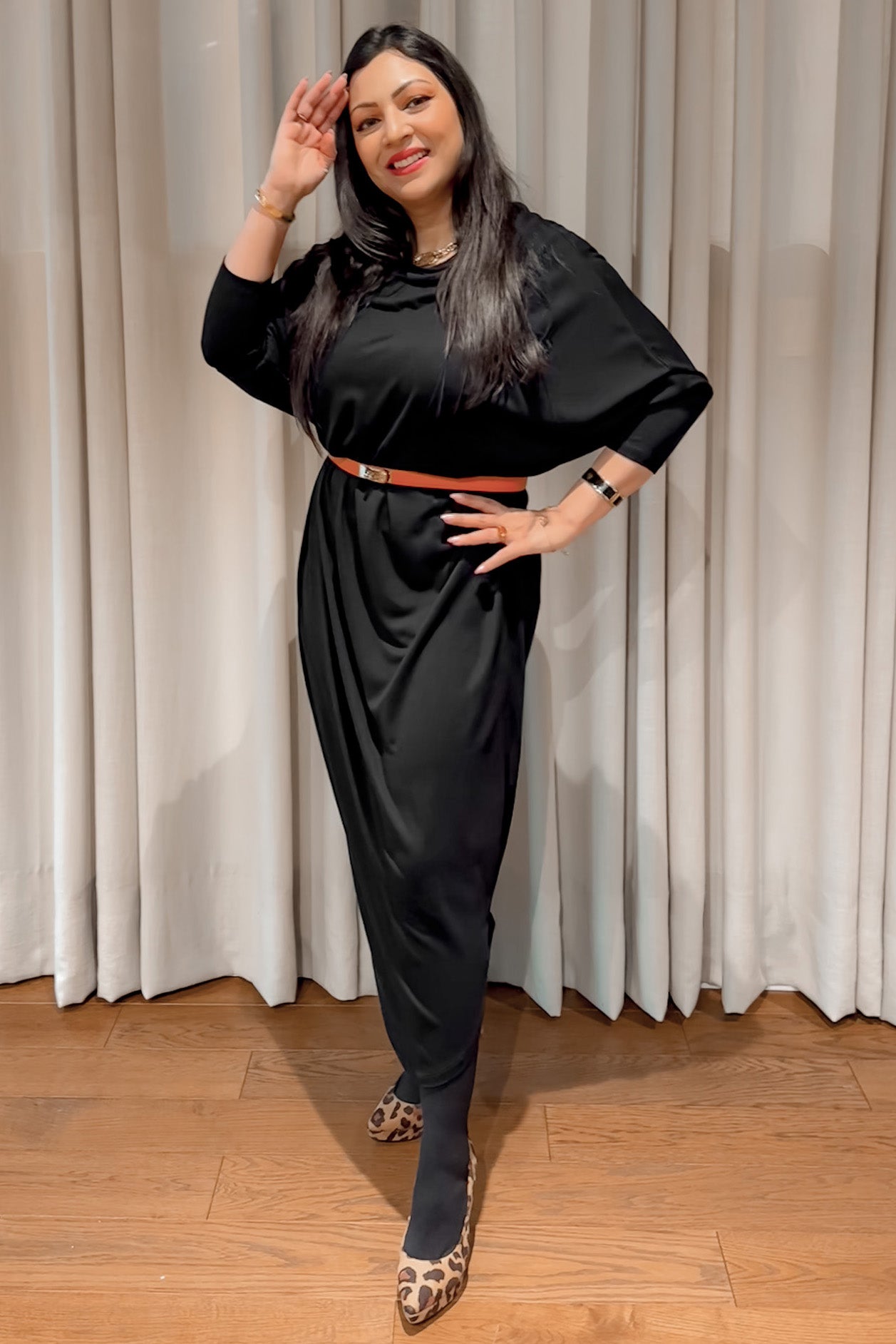 A woman poses in front of white curtains, wearing an AWAN Sweater Kaftan dress in warm black. She accessorizes with a brown belt and leopard print flats, smiling.